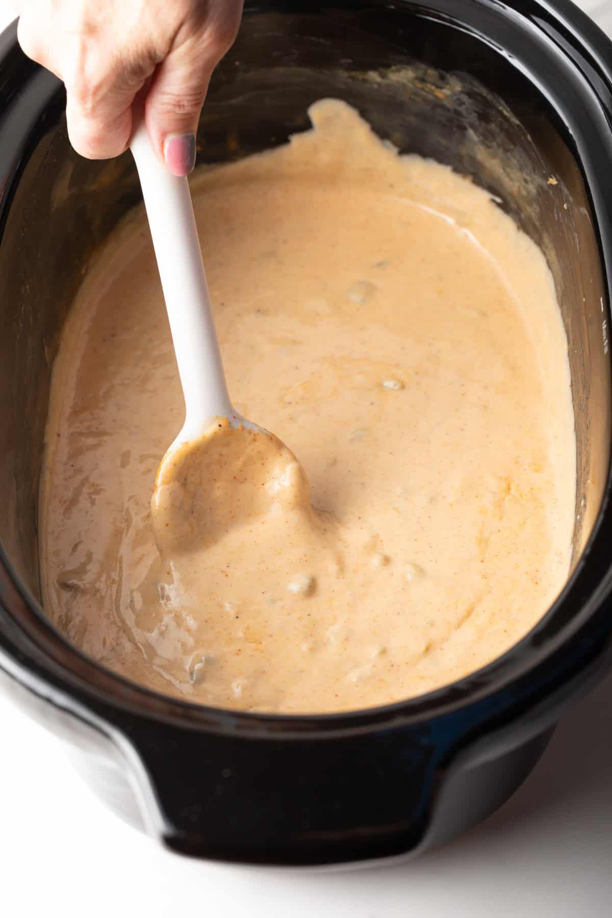 A white spoon stirring slow cooker queso dip in a black crock pot.