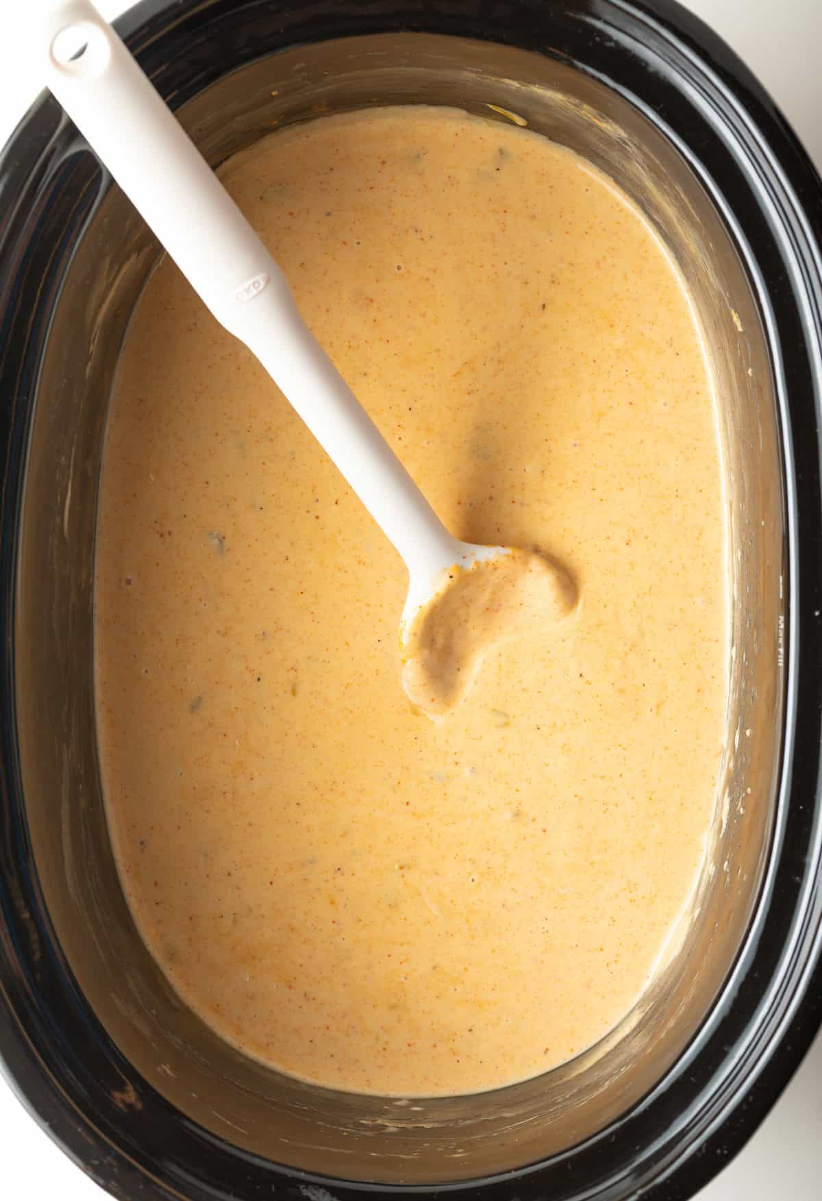 Black slow cooker filled with queso dip with a white spoon stirring it.
