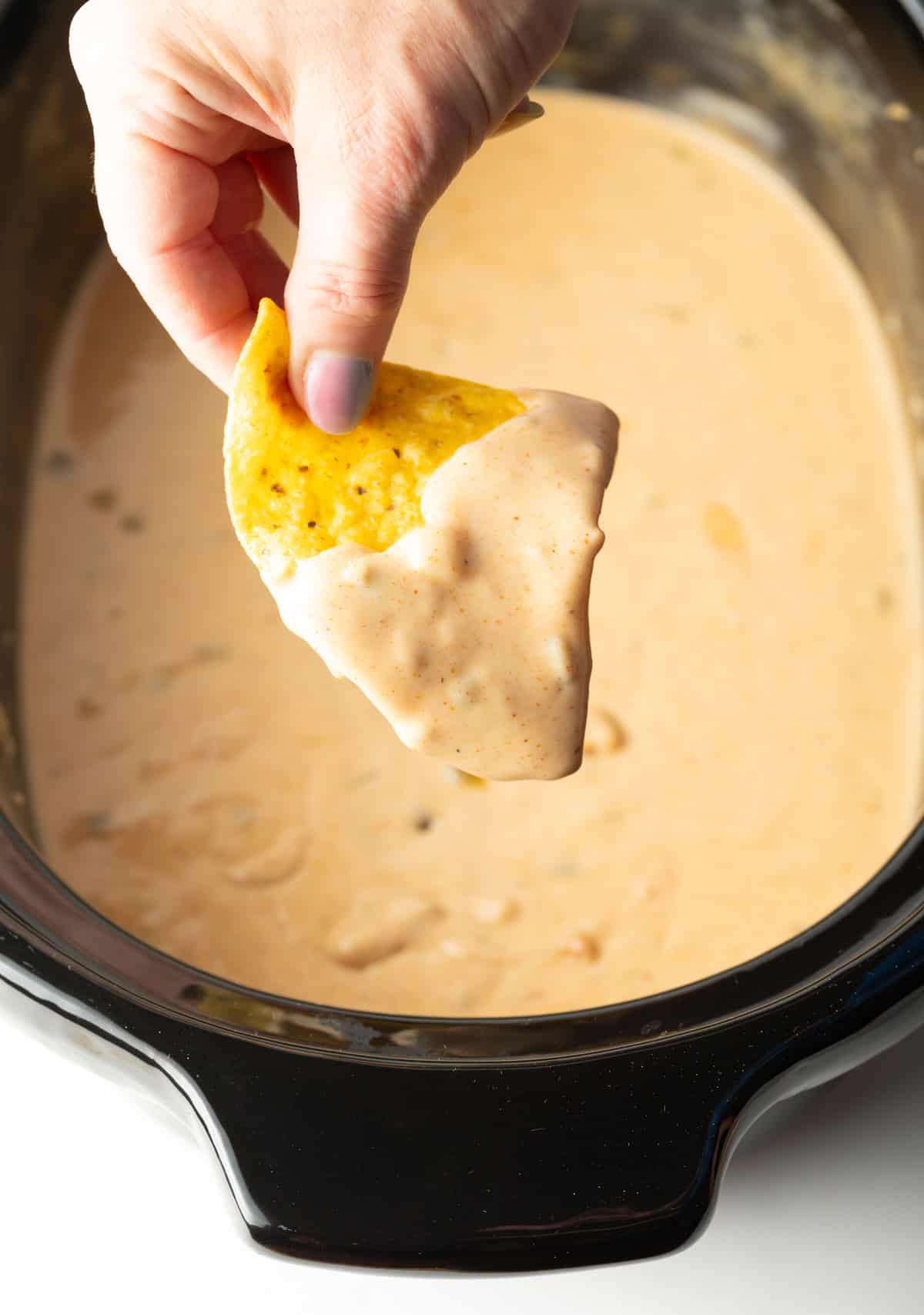 A hand holding a tortilla chip that's been dipped in crock pot queso hovered over the crock pot.