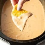 A hand holding a tortilla chip that's been dipped in crock pot queso dip, hovered over the crock pot.