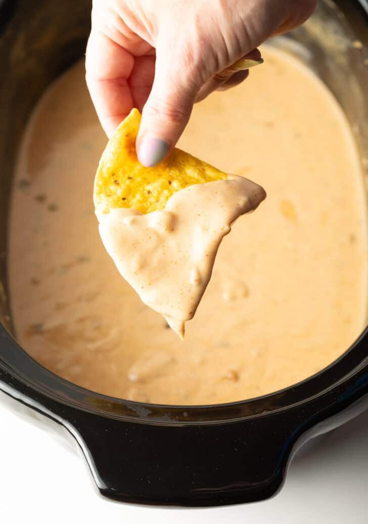 A hand holding a tortilla chip that's been dipped in crock pot queso dip, hovered over the crock pot.