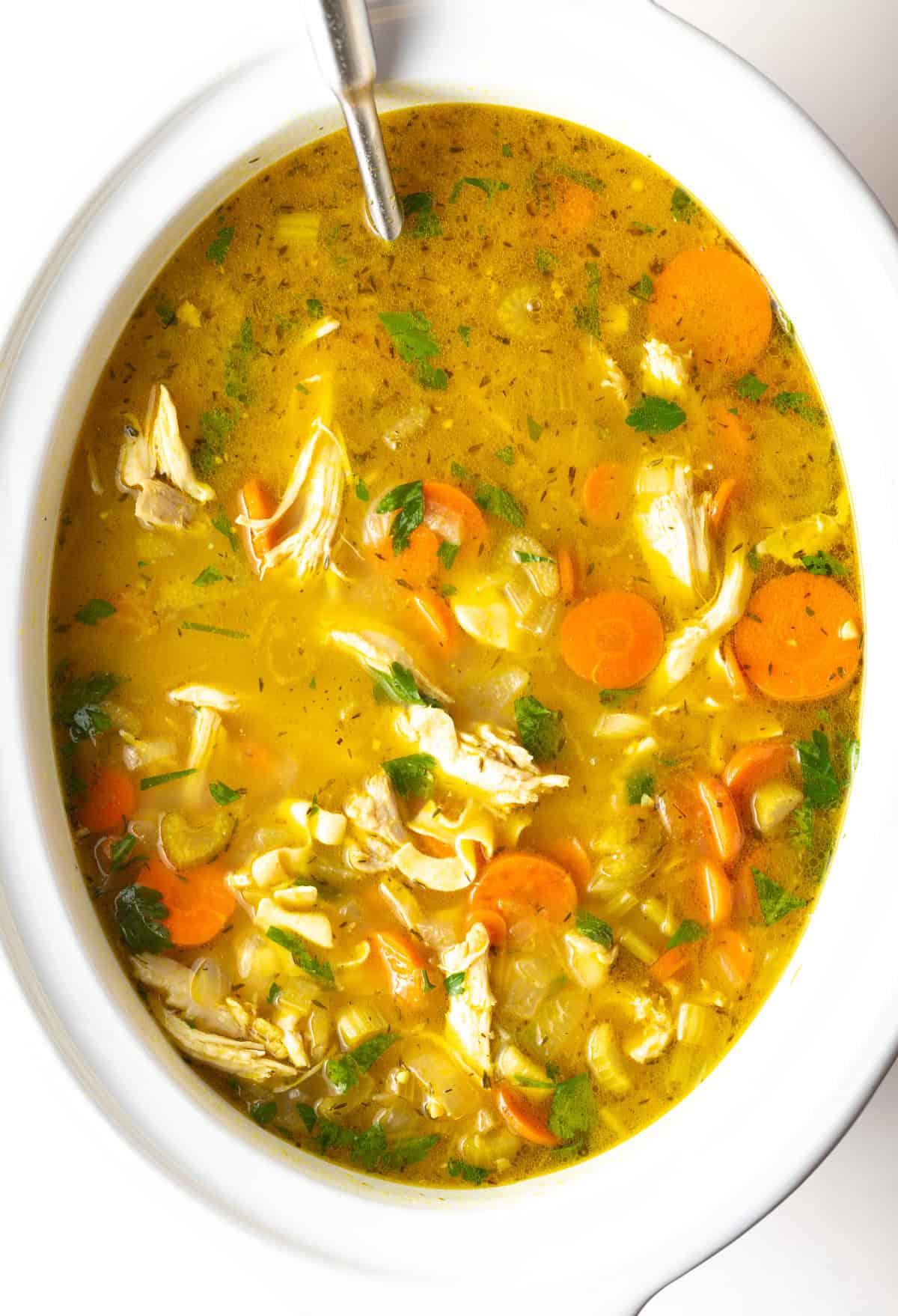 Chicken noodle soup in a white crockpot with a metal ladle.