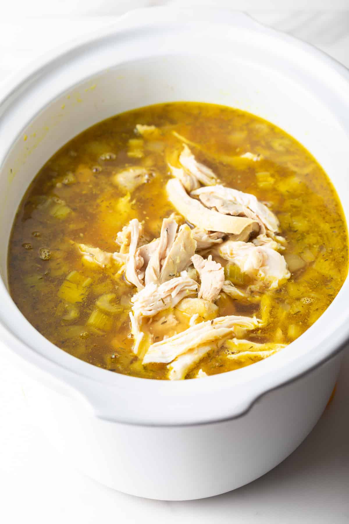 Shredded chicken being added to crockpot chicken noodle soup in a white slow cooker.