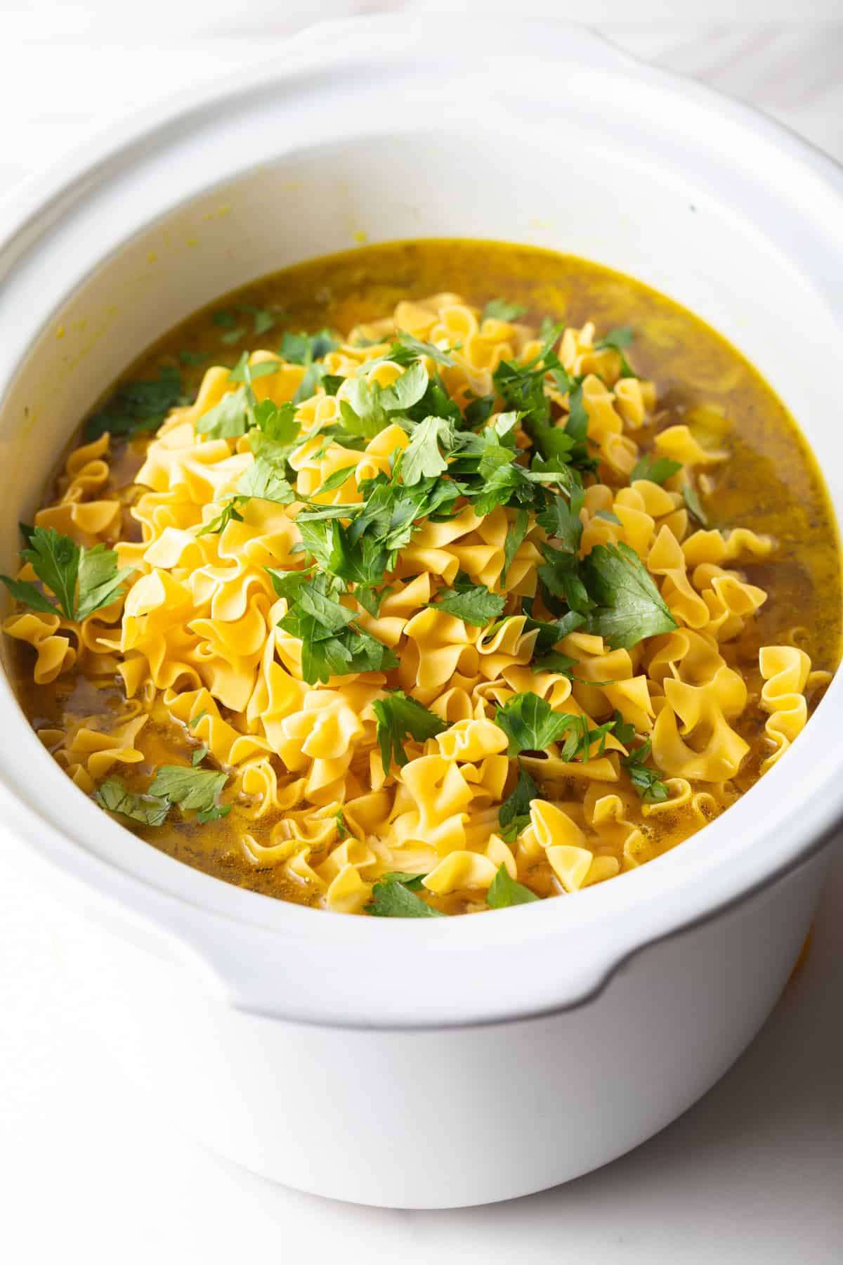 Uncooked egg noodles and parsley are being added to the broth mixture for slow cooker chicken noodle soup. 