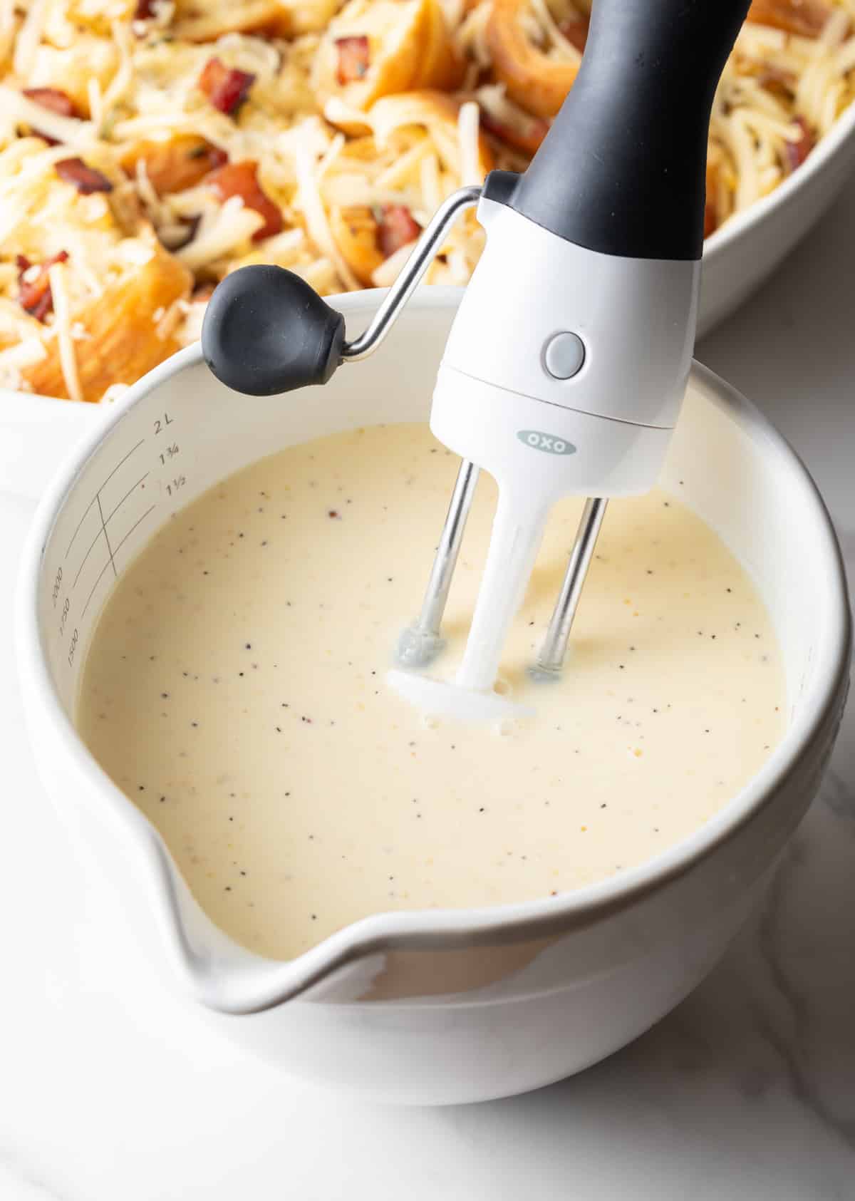 Cream, eggs, and mustard are being mixed together with a hand mixer in a white mixing bowl.