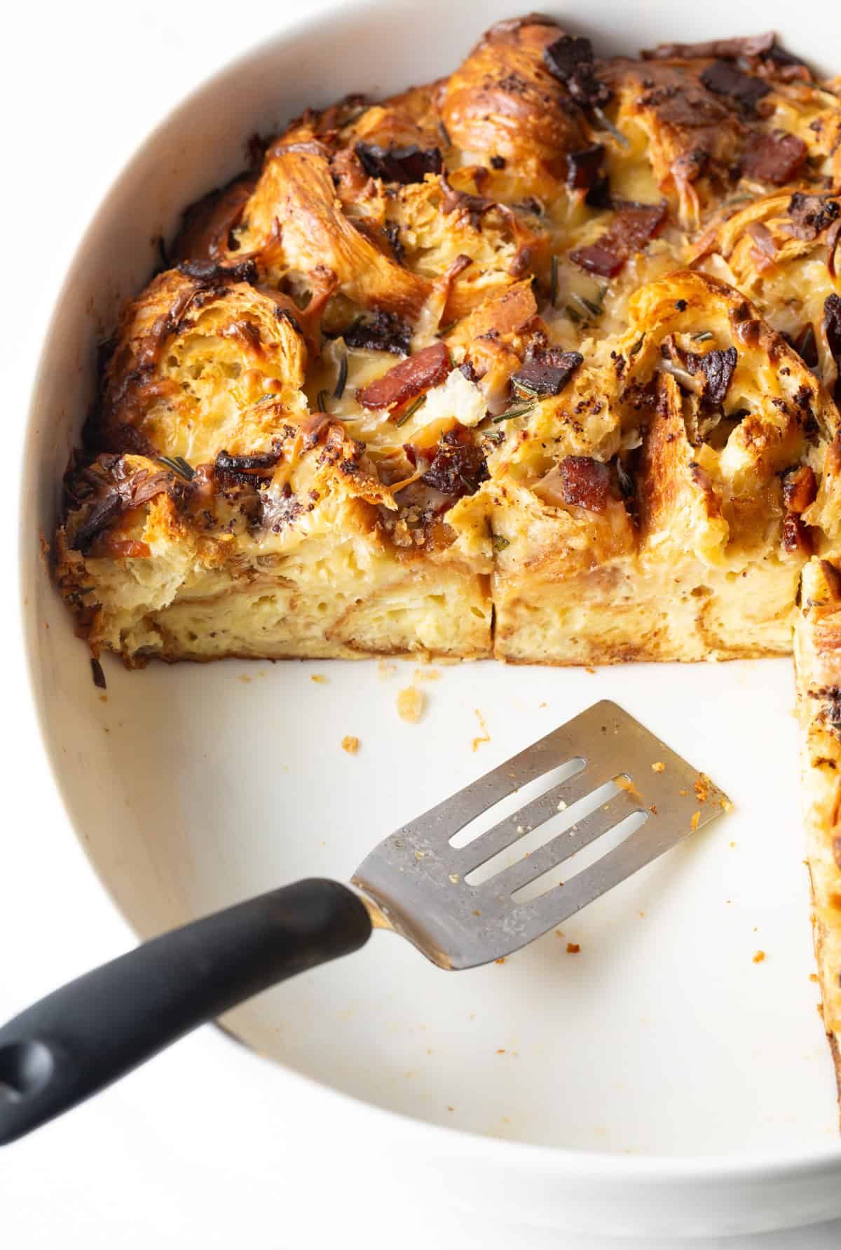 A metal spatula is serving slices of croissant casserole from a white baking dish.