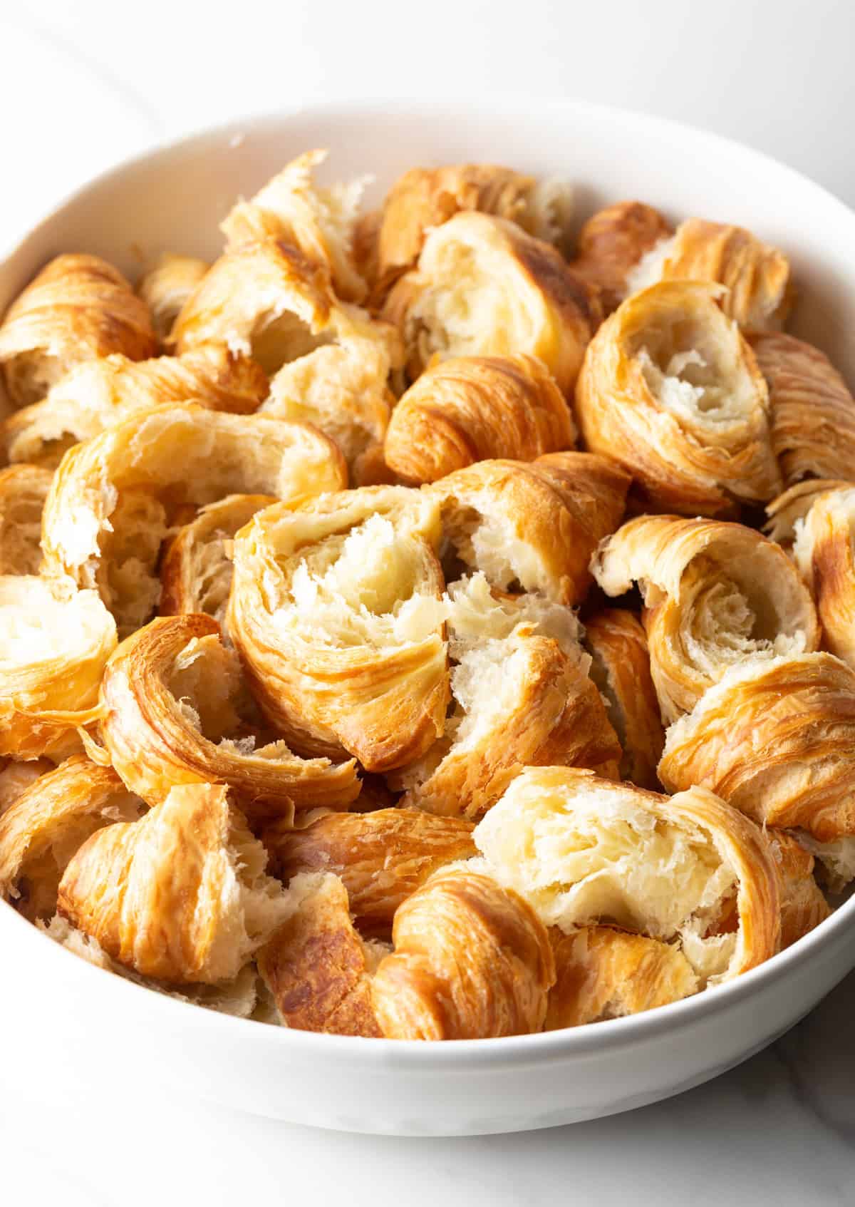 Croissants are torn into chunks and layered in a white casserole dish.