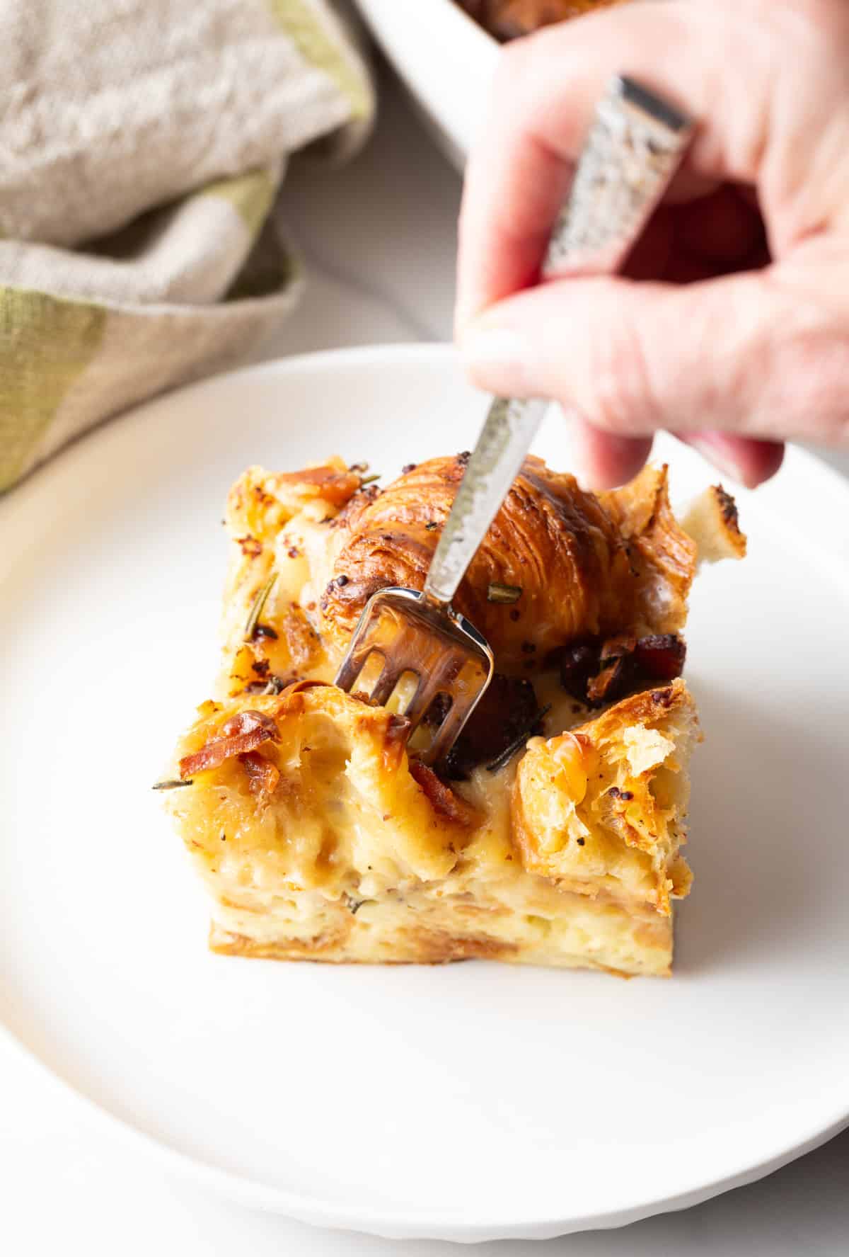 A fork is taking a bite out of a slice of croissant casserole that is sitting on a white plate.