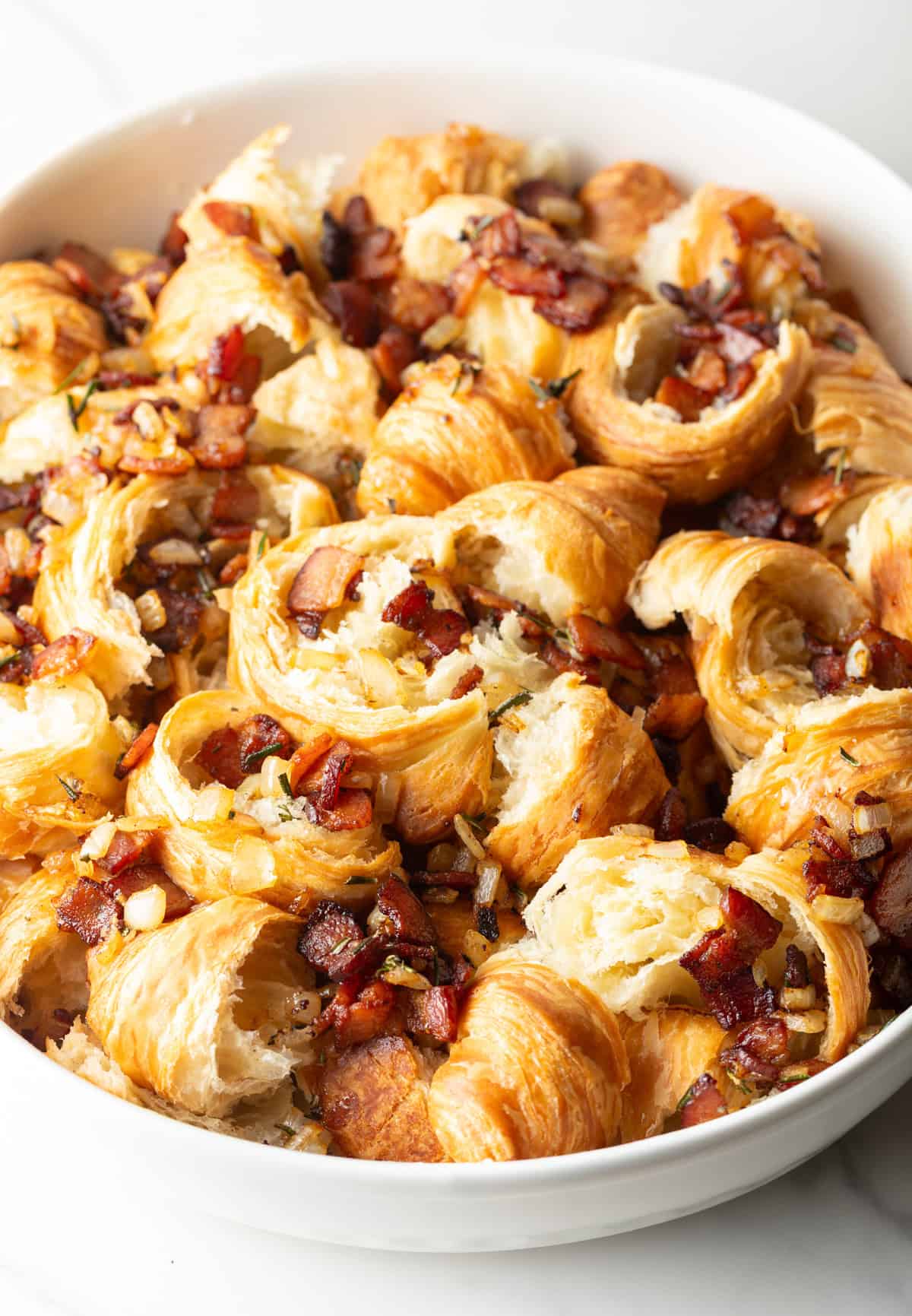 A bacon and vegetable mixture is being added to torn croissants in a white baking dish. 