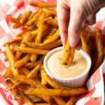 A basket of crispy French fries with a ramekin of French fry dipping sauce, and a hand is dipping two fries into the sauce.