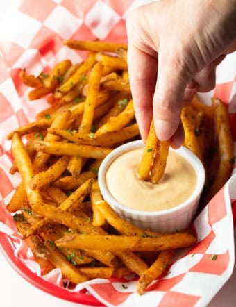A basket of crispy French fries with a ramekin of French fry dipping sauce, and a hand is dipping two fries into the sauce.