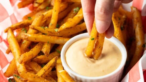 A basket of crispy French fries with a ramekin of French fry dipping sauce, and a hand is dipping two fries into the sauce.