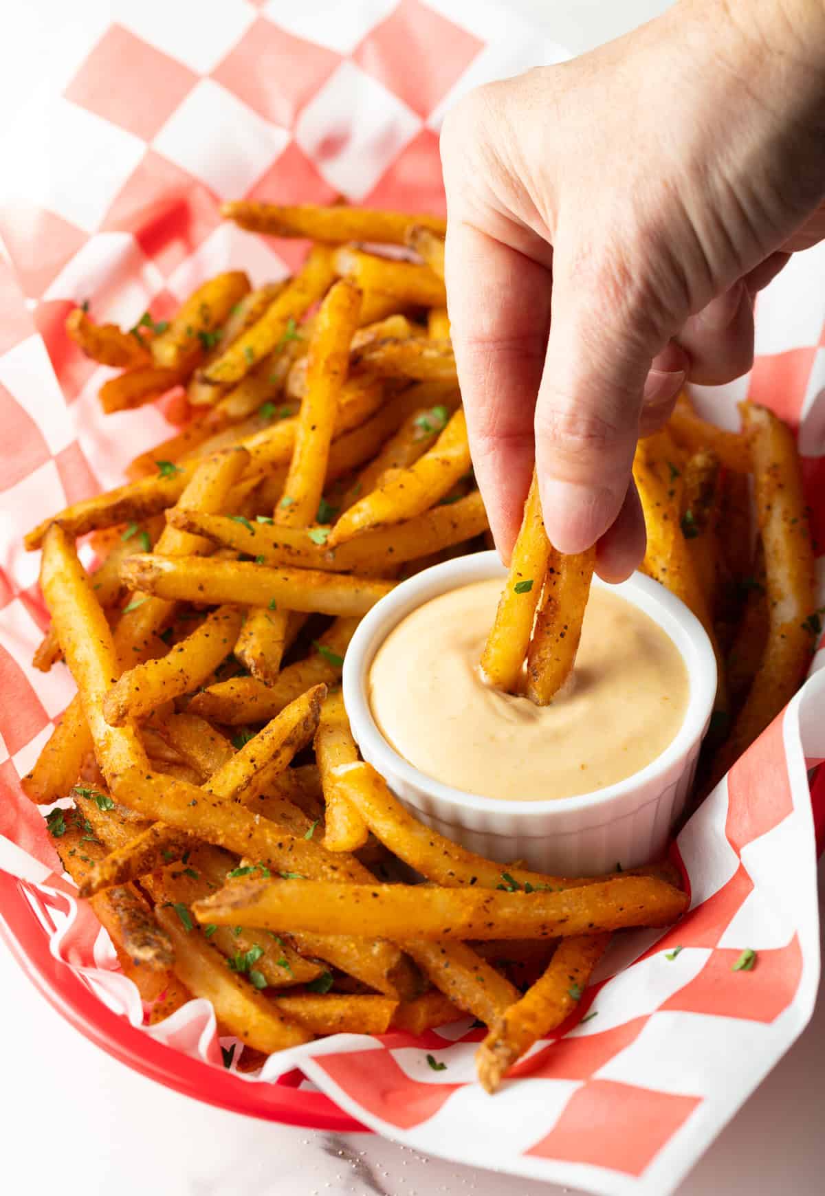 A basket of crispy French fries with a ramekin of French fry dipping sauce, and a hand is dipping two fries into the sauce.