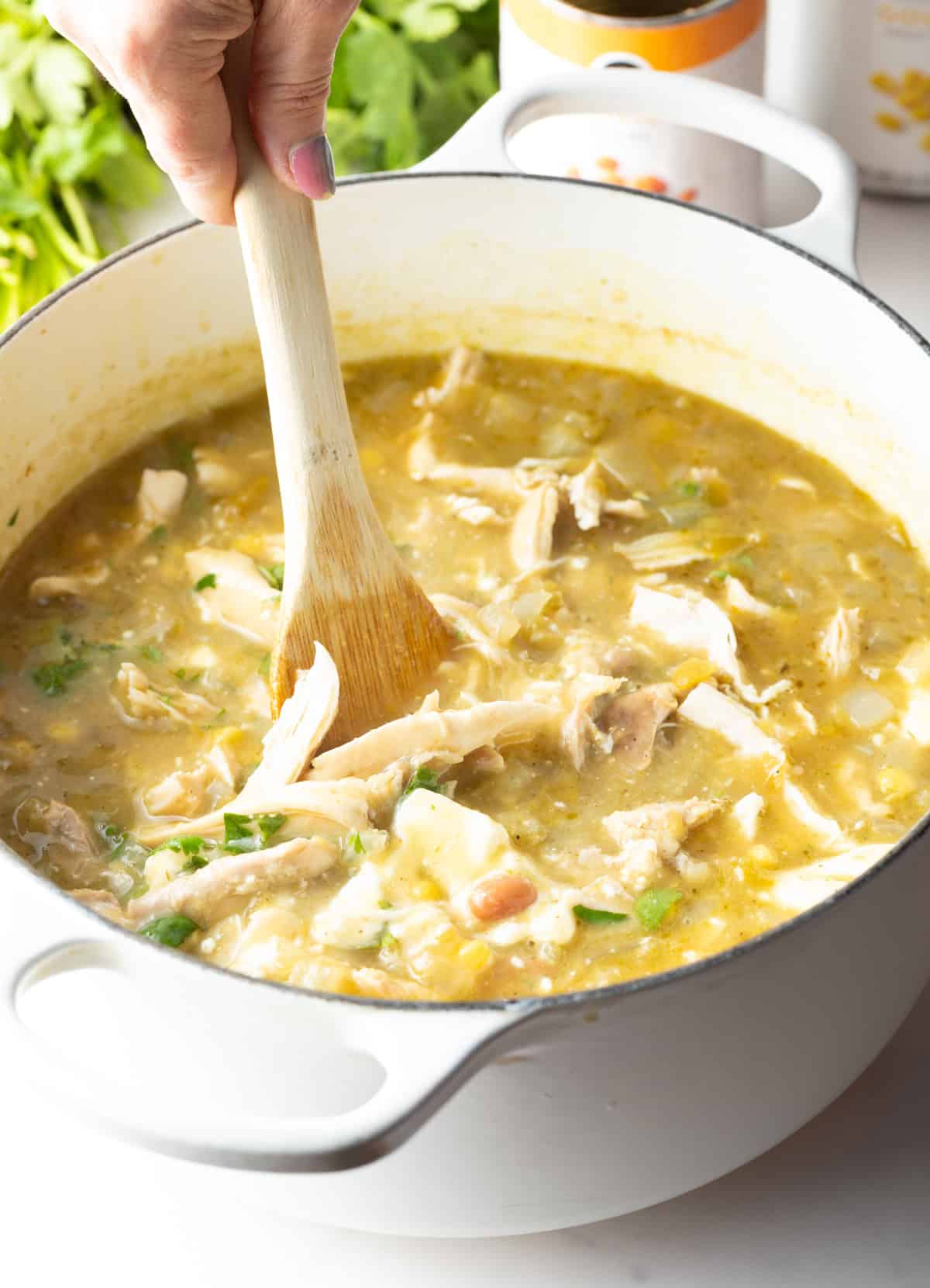 Hand using a large wooden spoon to stir shredded chicken into a large white cooking pot.