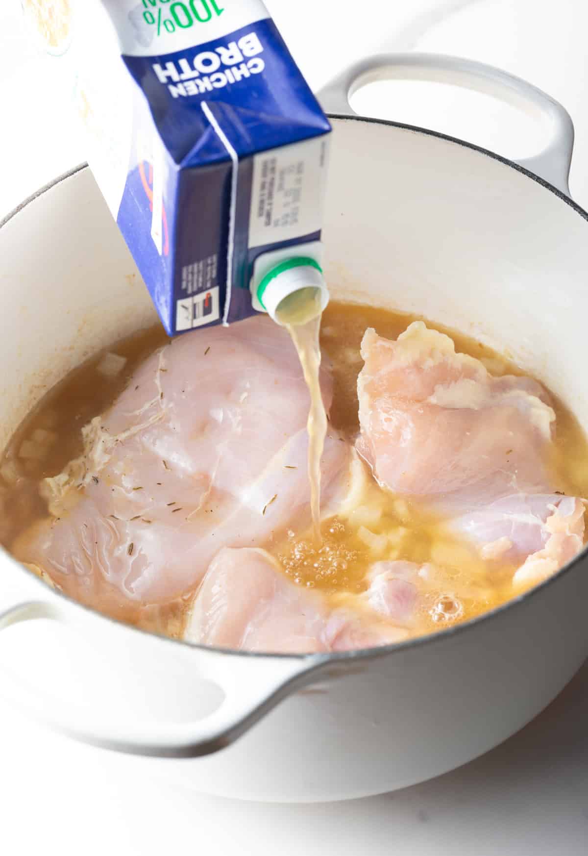 Adding chicken broth from a carton to a soup pot with raw chicken breasts.