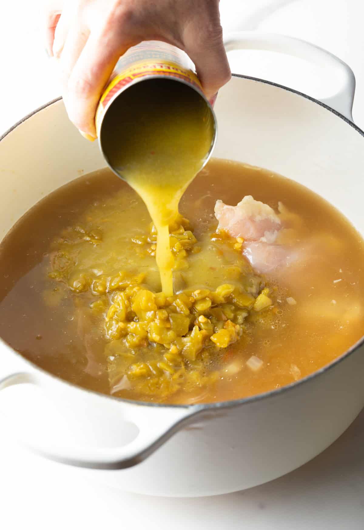Pouring verde enchilada sauce from a can into a white pot of soup.