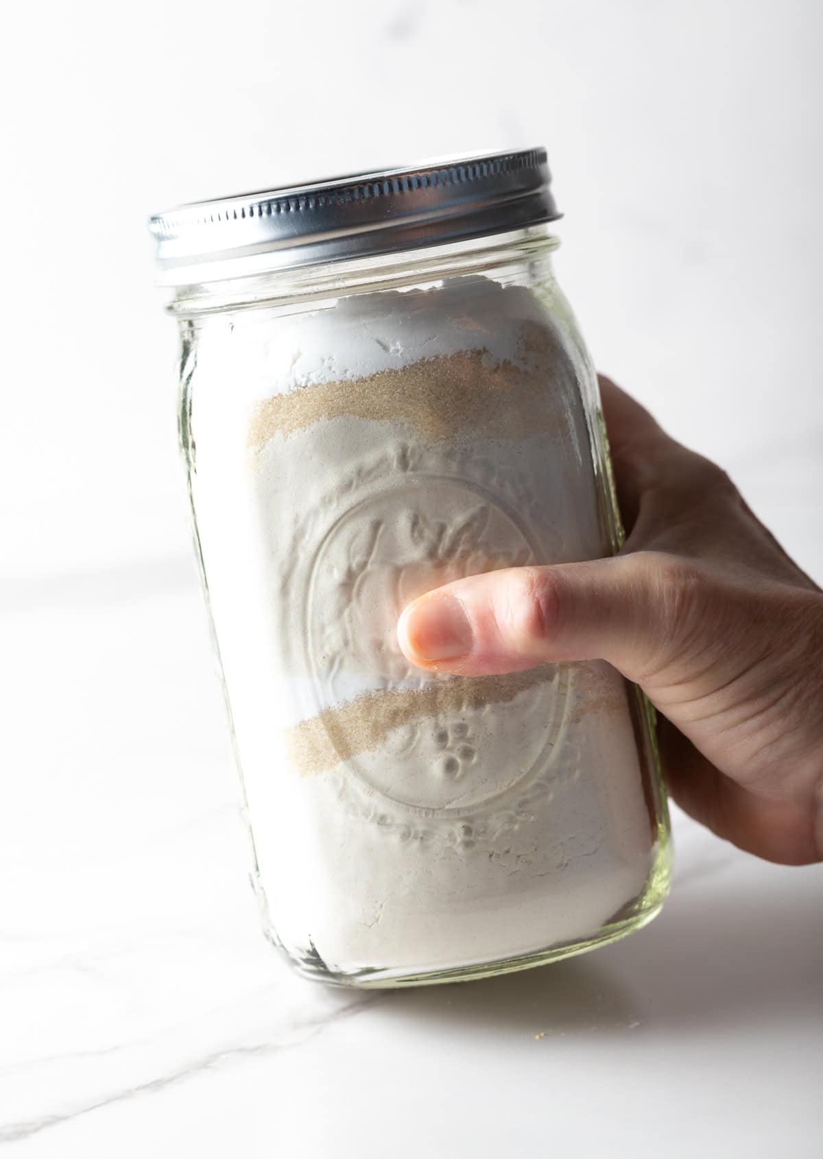 A hand holding a glass jar filled with homemade pancake mix ingredients, ready to be shaken.