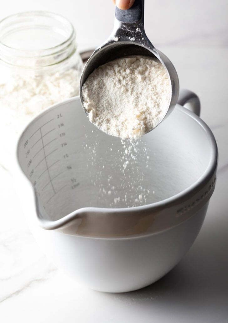 A scoop of homemade pancake mix is being poured into a mixing bowl.