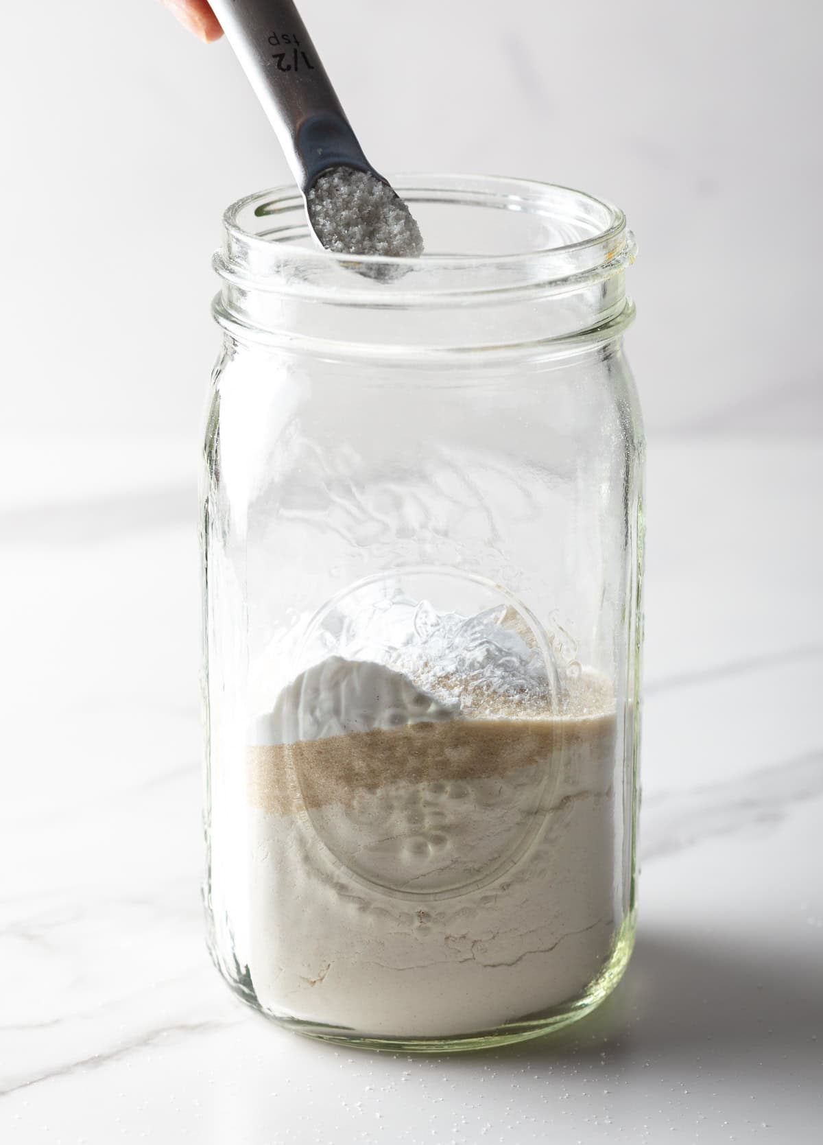 A measuring spoon scooping baking soda into a glass jar of homemade pancake mix.