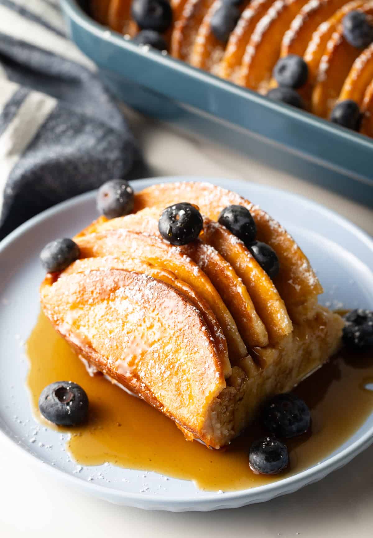 A serving of easy pancake breakfast casserole on a blue plate, surrounded by syrup and blueberries.