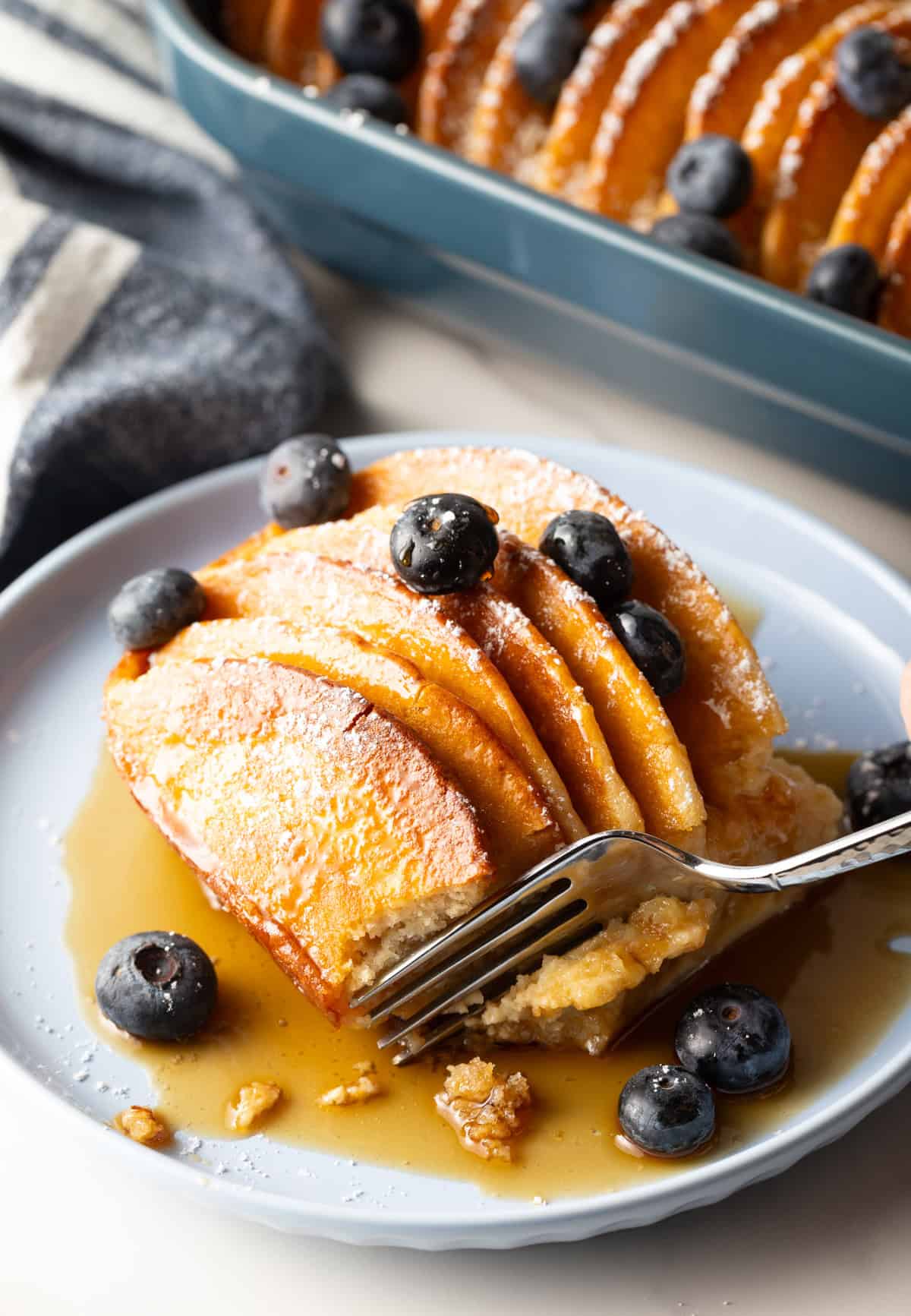 A serving of easy pancake casserole on a blue plate, surrounded by syrup and blueberries, with a fork cutting a bite.