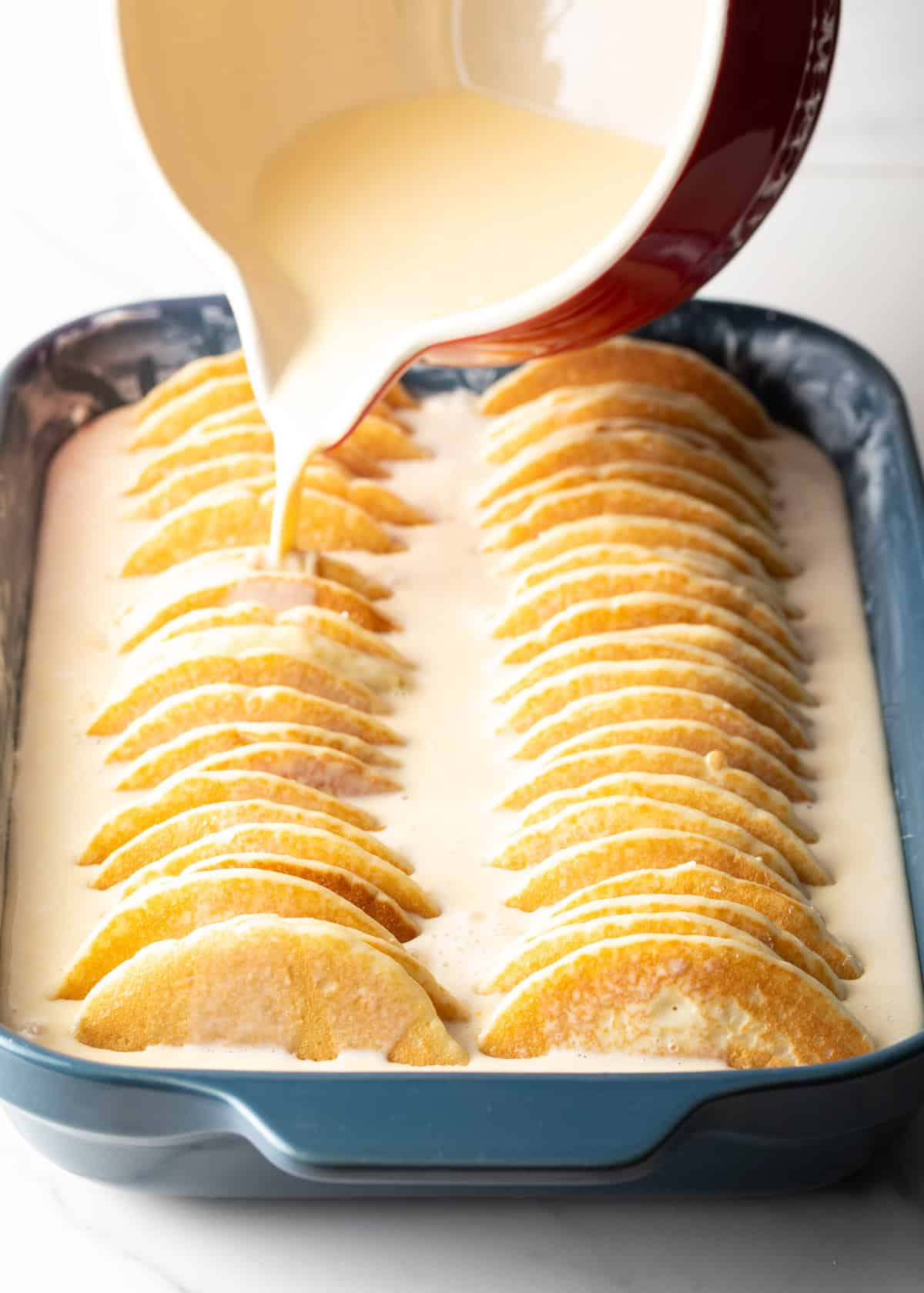Cream mixture is being poured over an un-baked pancake casserole in a baking dish. 