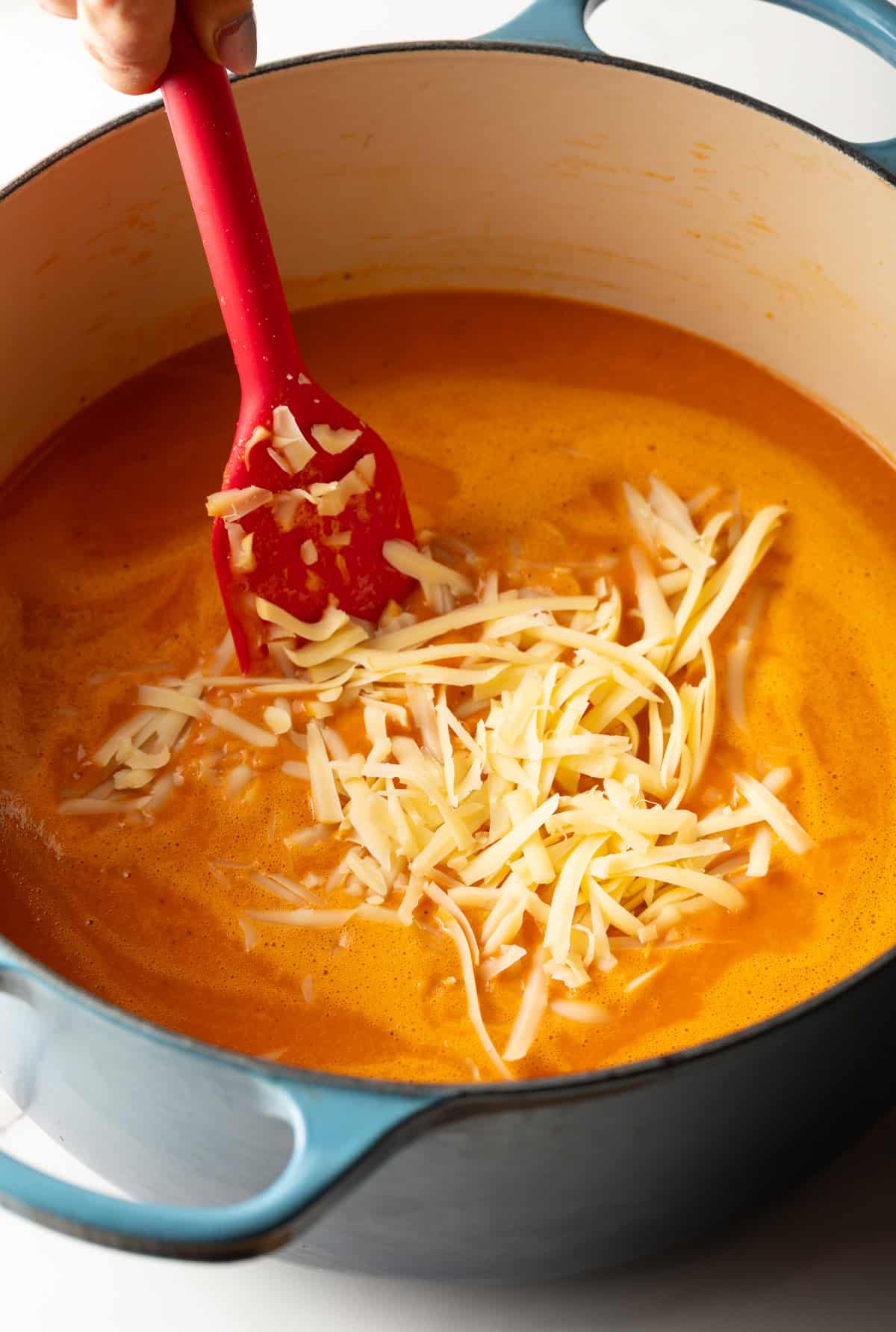 Shredded gouda is being stirred into a pot of red pepper soup.