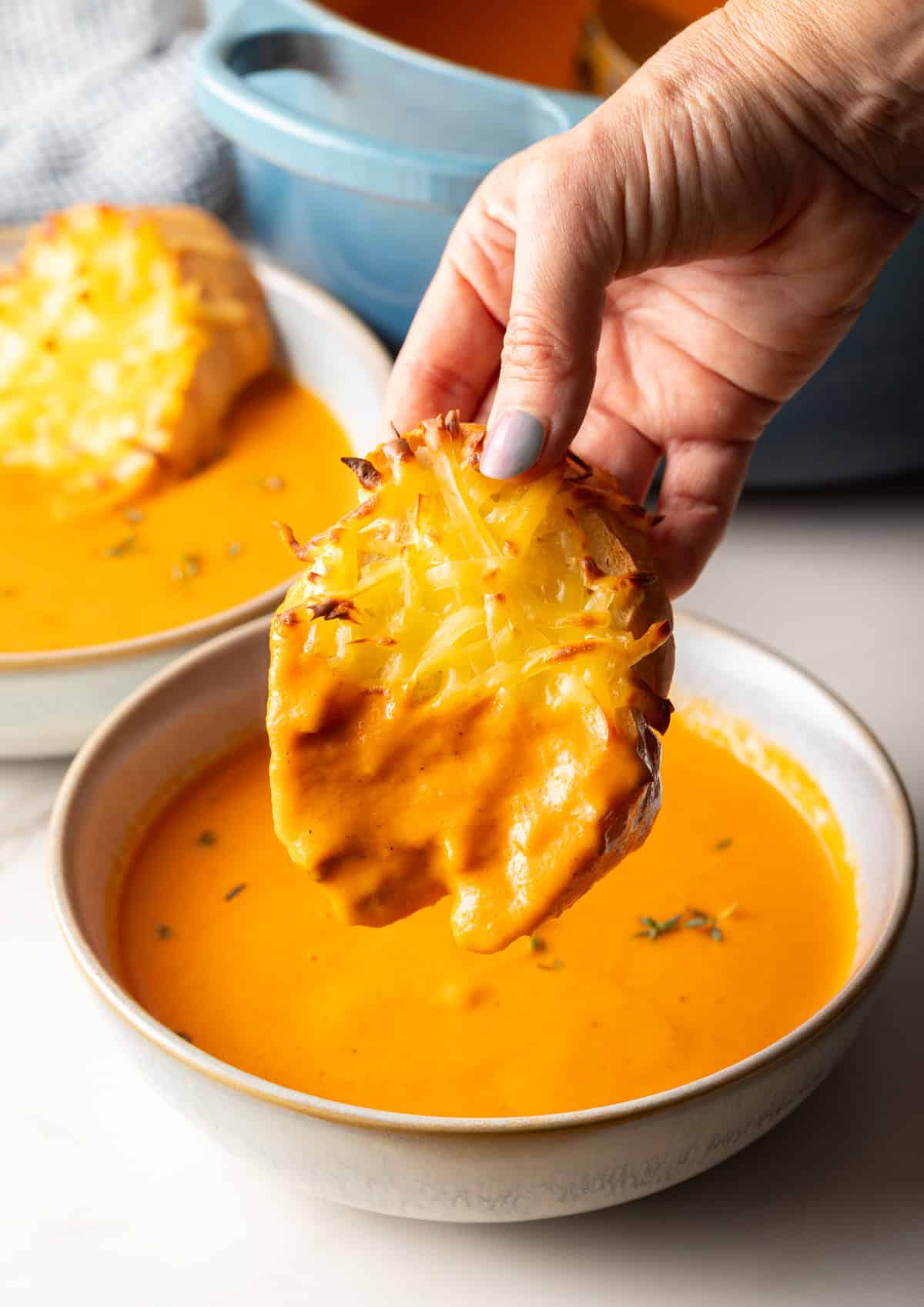A hand holding cheesy bread that is being dipped into a bowl of roasted red pepper soup with gouda.