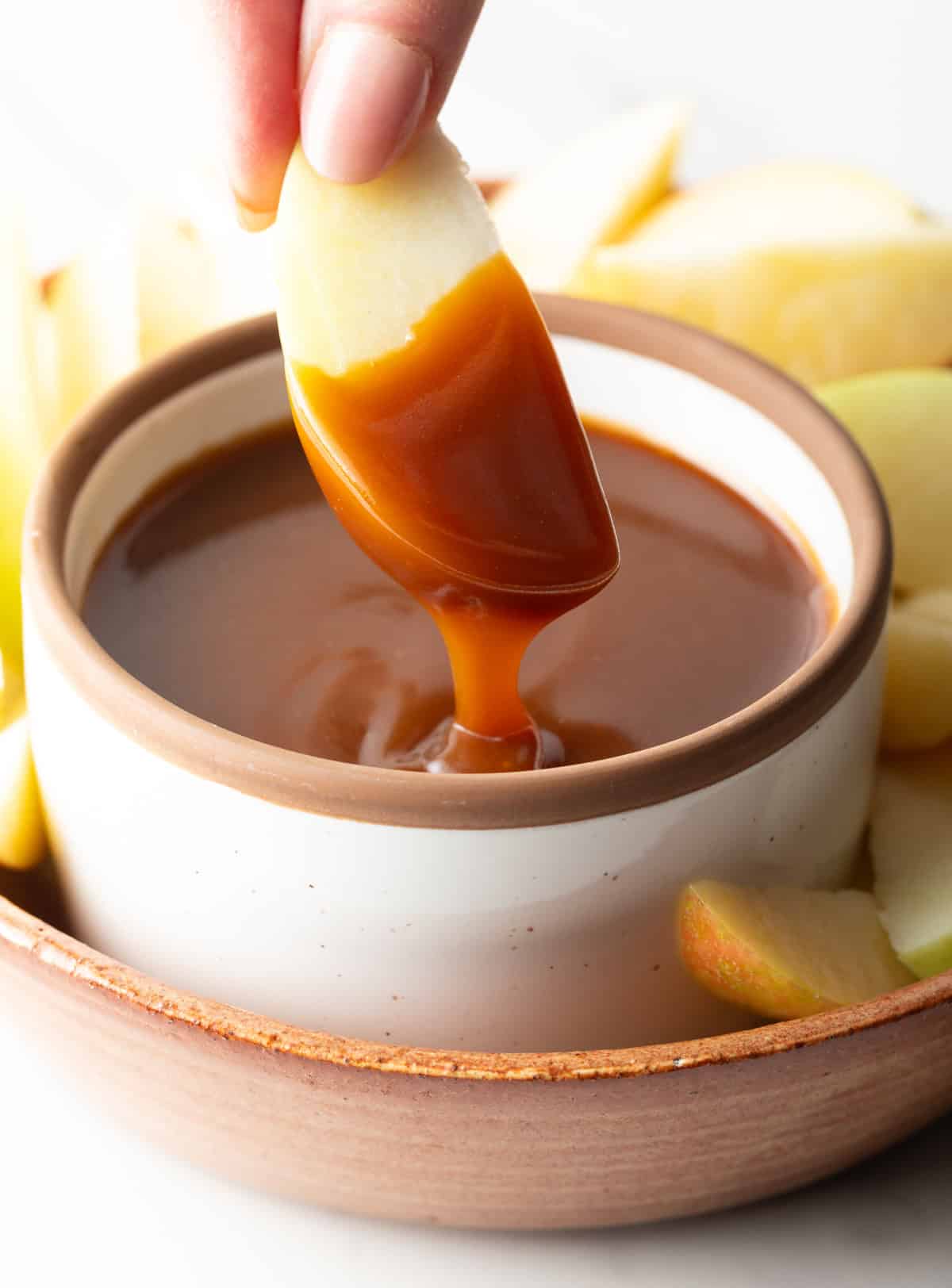 An apple sliced that's been dipped into salted caramel sauce hovering over a jar of sauce.