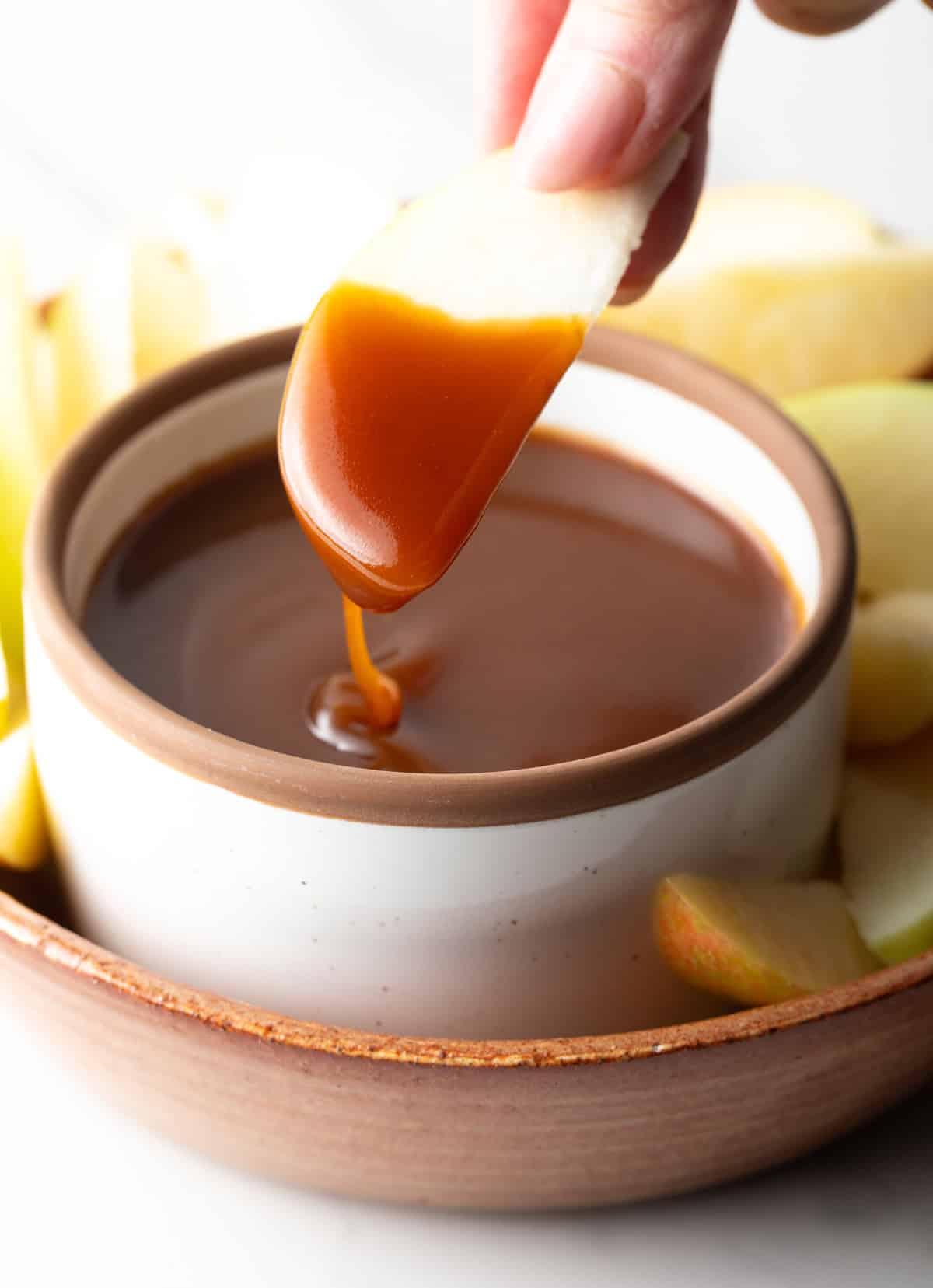 An apple slice being dipped into a container of salted caramel sauce.