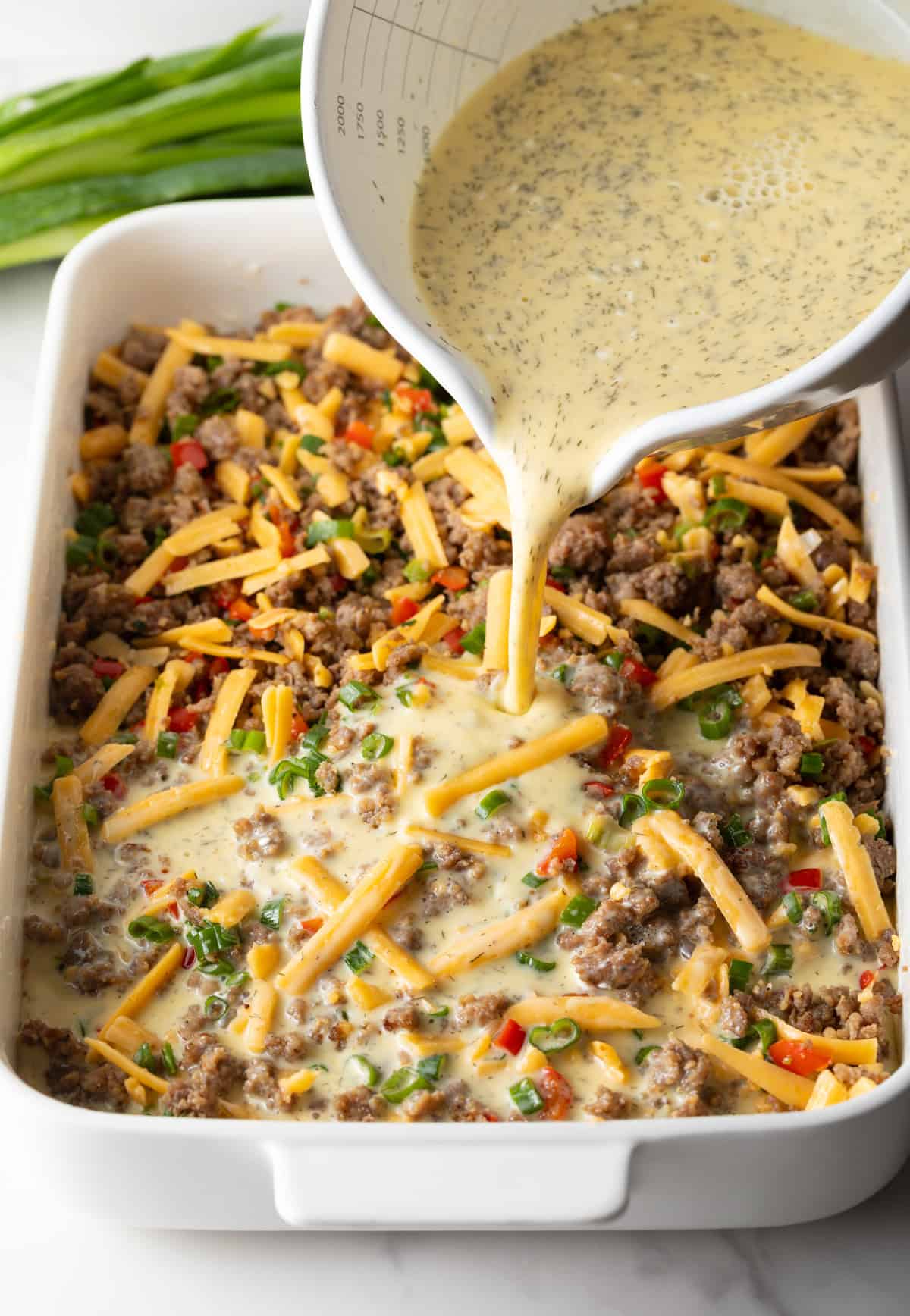 Pouring egg mixture over shredded cheese and sausage in a white rectangle baking dish.