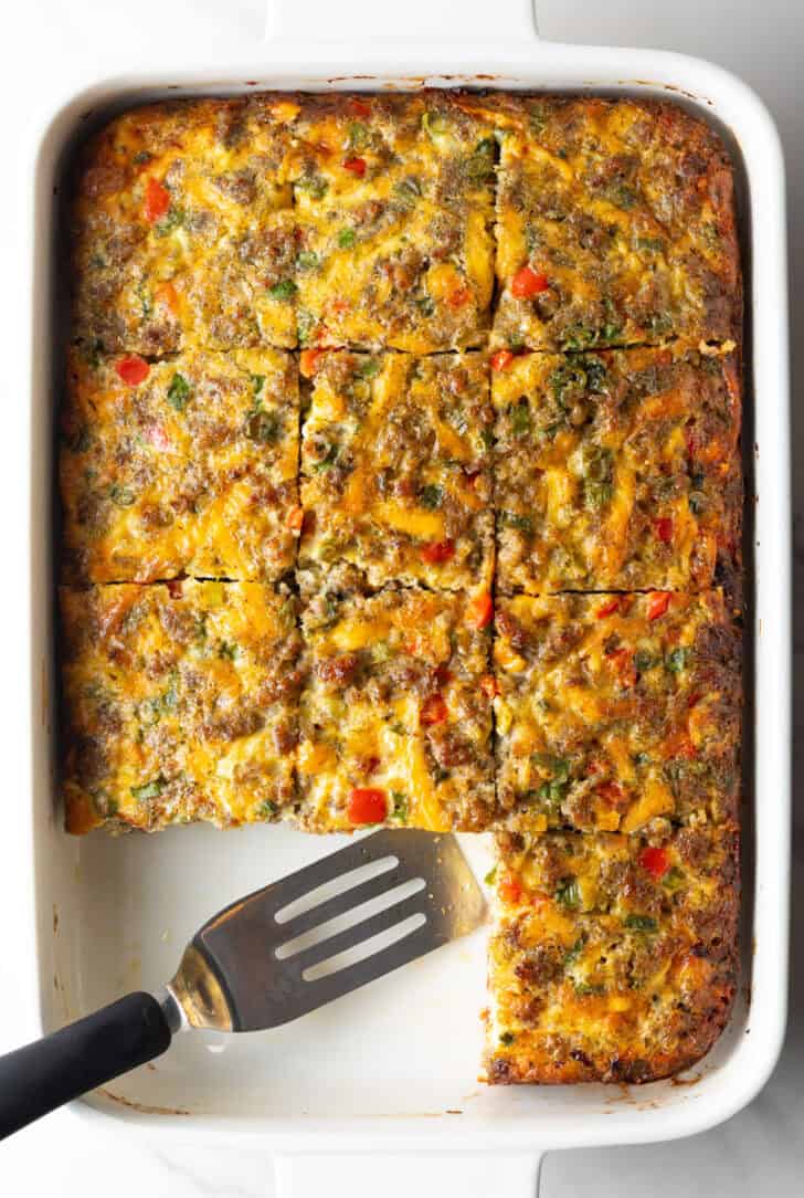 Pan of egg casserole with sausage and vegges. A spatula is in the pan, with square portions of the casserole removed.