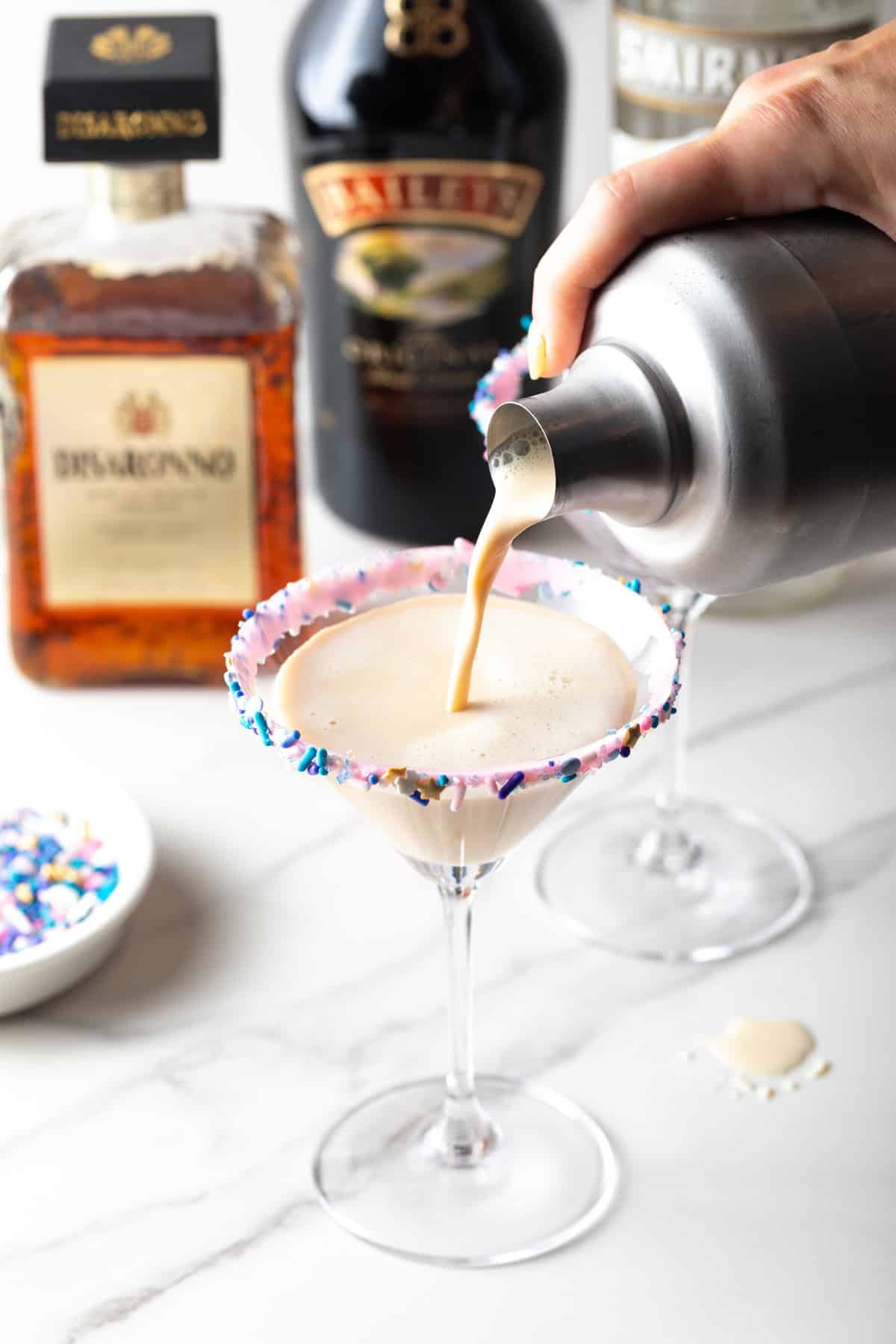Creamy sugar cookie cocktail with a colorful rim, surrounded by liquor bottles.