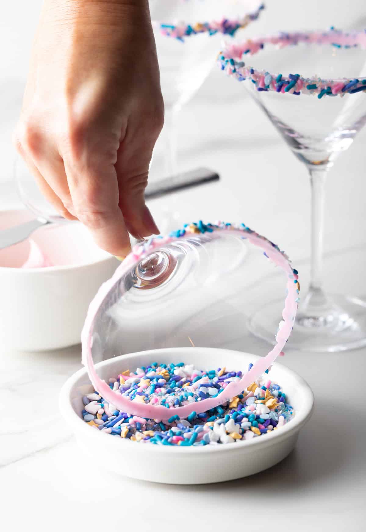 A martini glass being dipped into colorful sprinkles to line the rim.