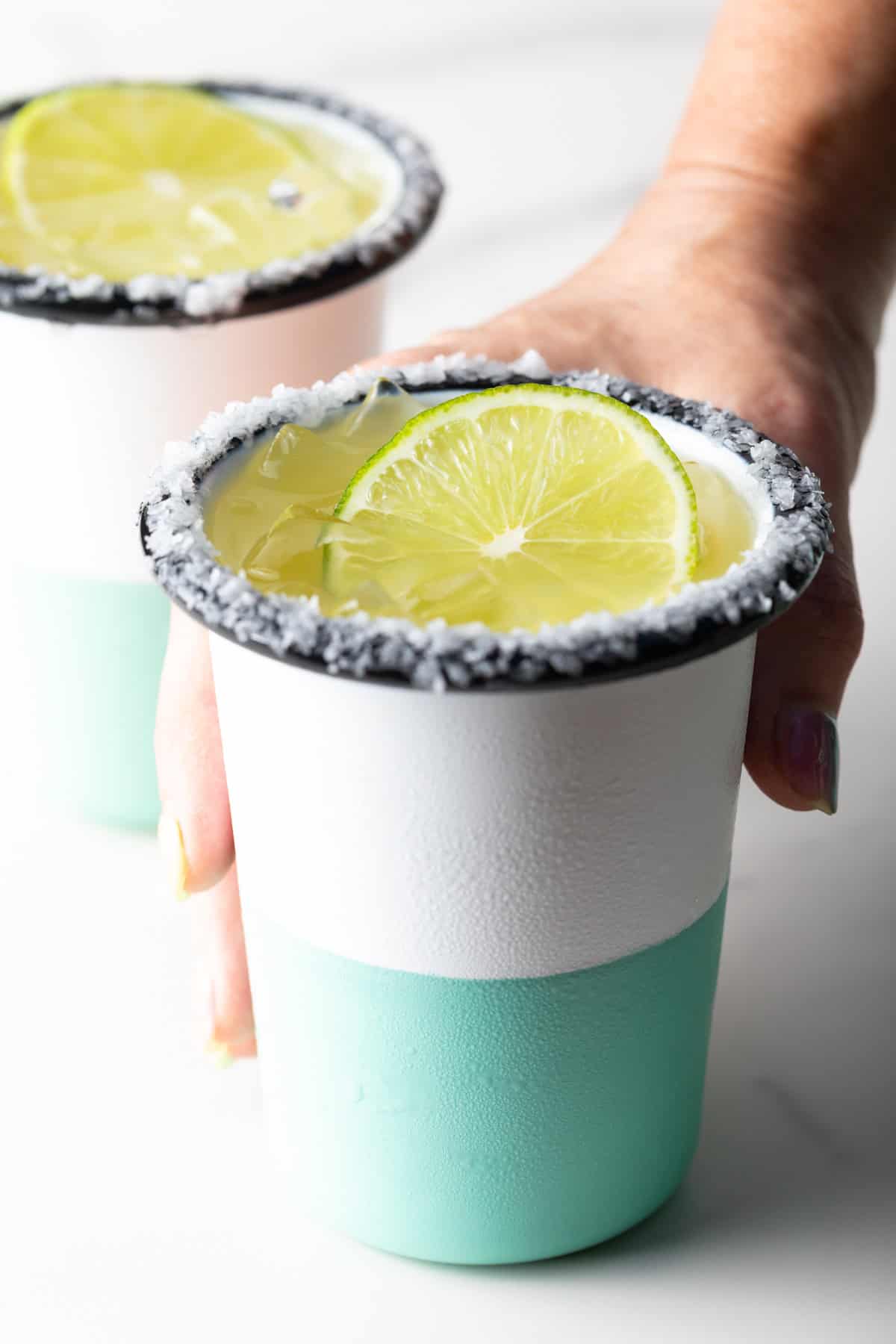 Hand grabbing a white and green-striped cup with salt around the rim and slice of lime in the drink.