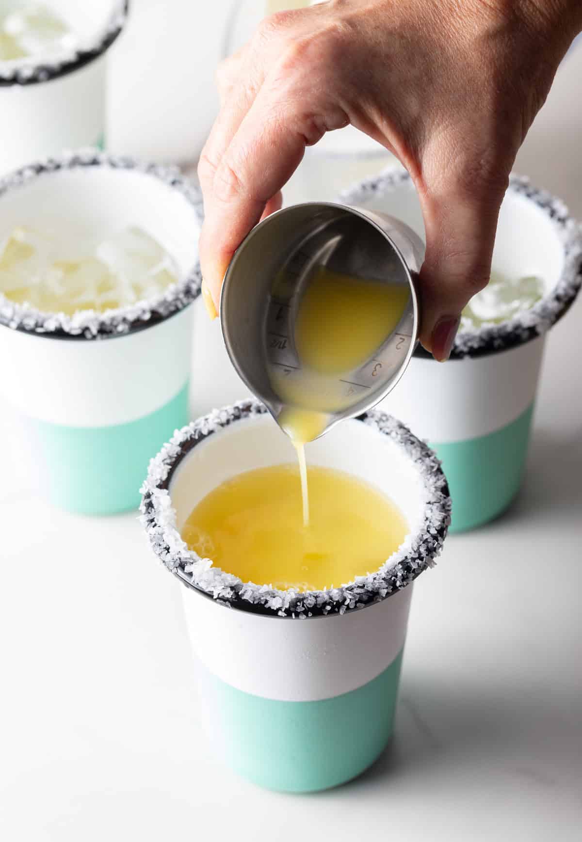 Hand pouring juice from a metal shot glass into a cup with a salted rim.