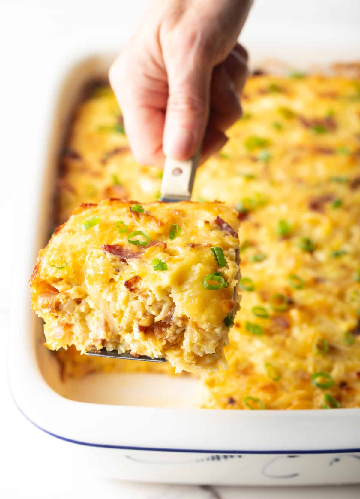 Hand using a metal spatula to lift a square of Amish breakfast casserole from a white baking dish.