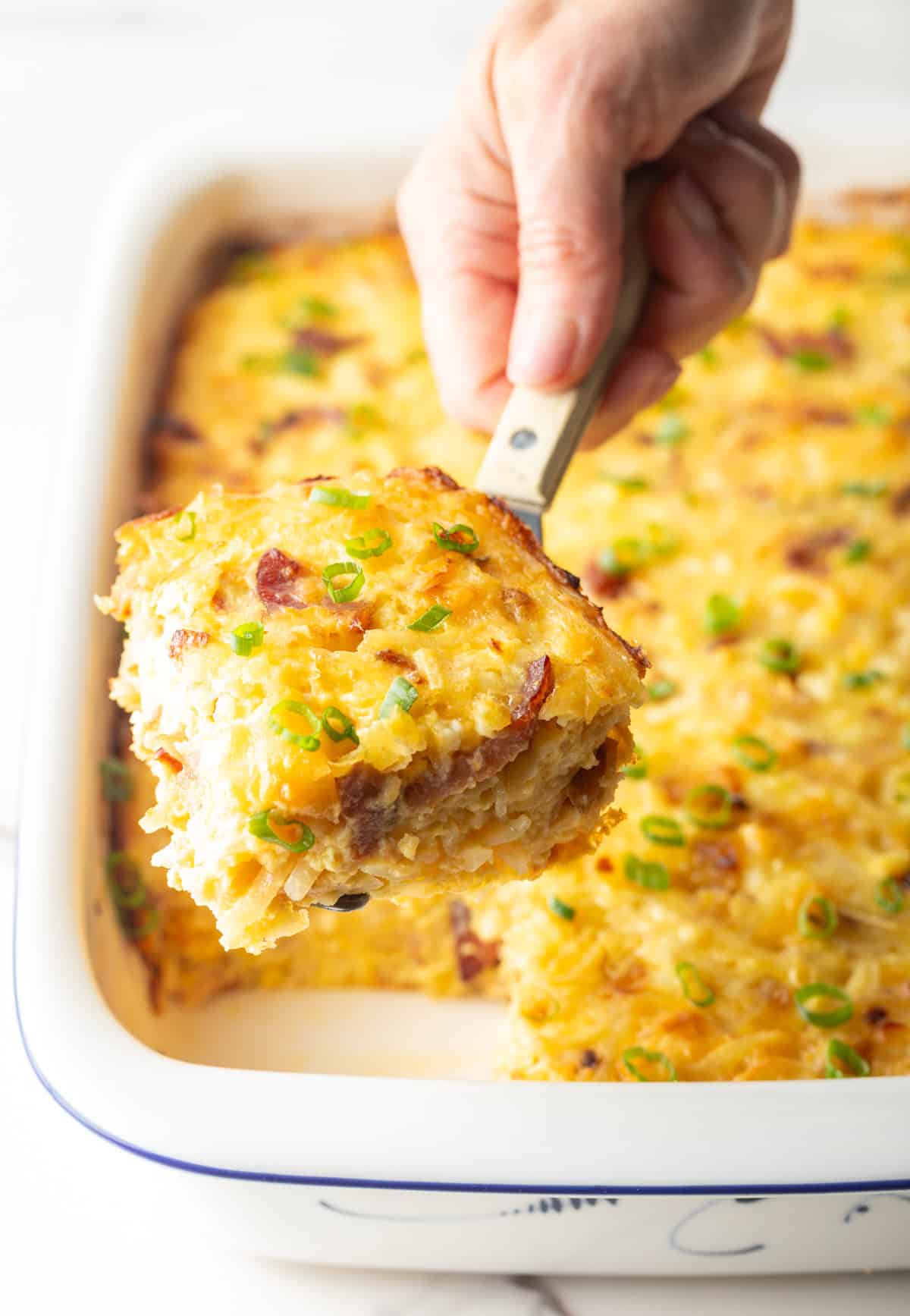 Hand using a metal spatula to lift a square of Amish breakfast casserole from a white baking dish.