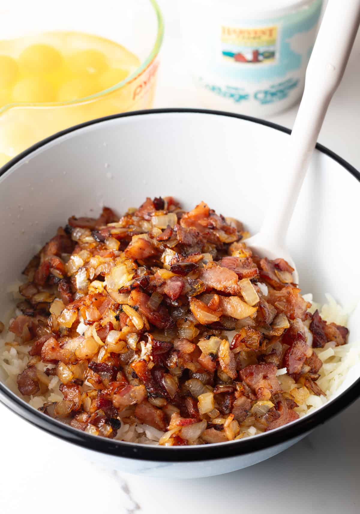 Adding cooked chopped bacon to a white mixing bow with shredded potatoes.
