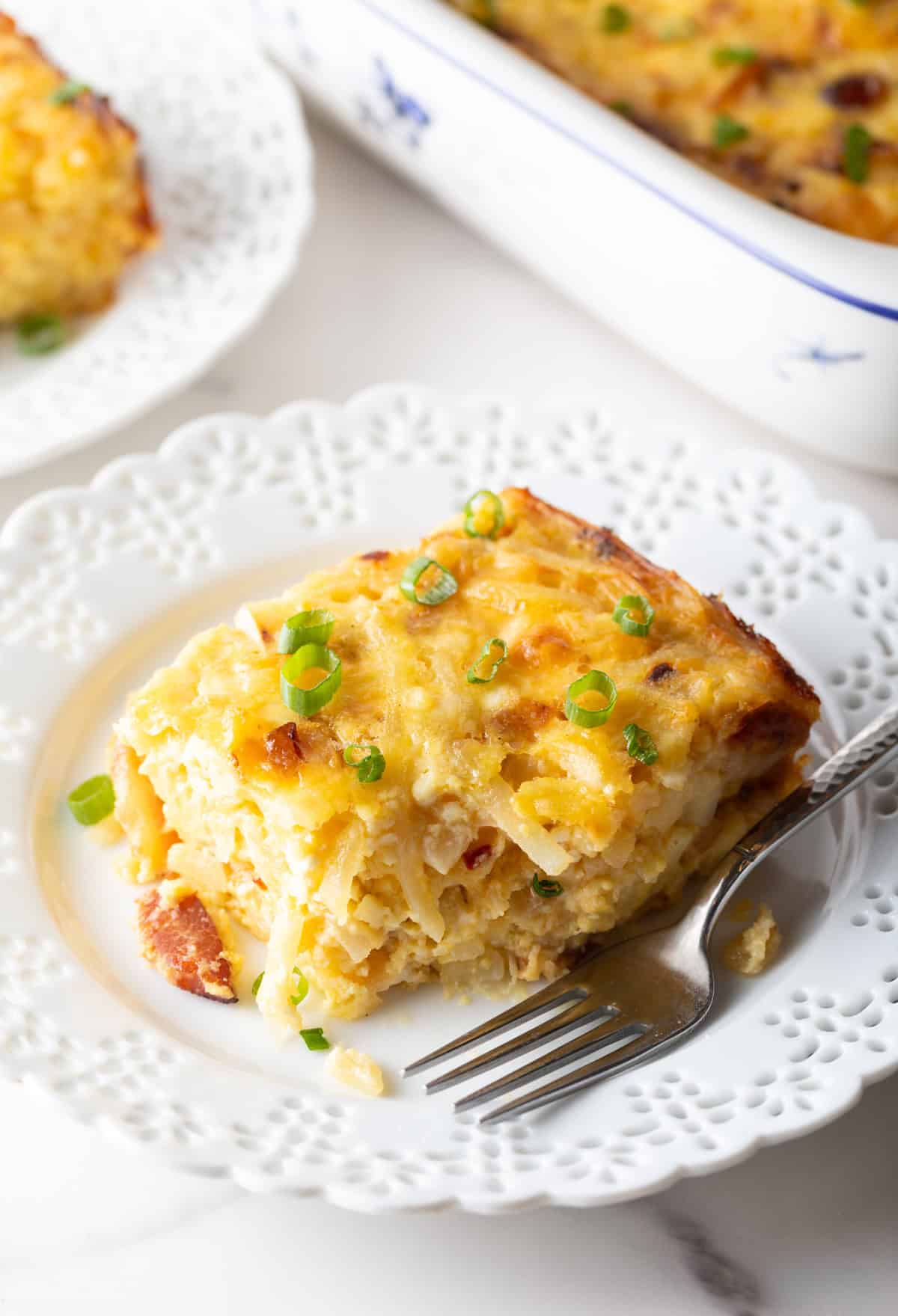 Square serving of hash brown potato, egg and cheese casserole on a white plate with a fork.
