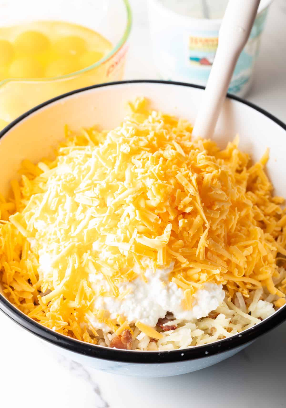 Adding shredded cheese to a bowl with cottage cheese, shredded potatoes and bacon.