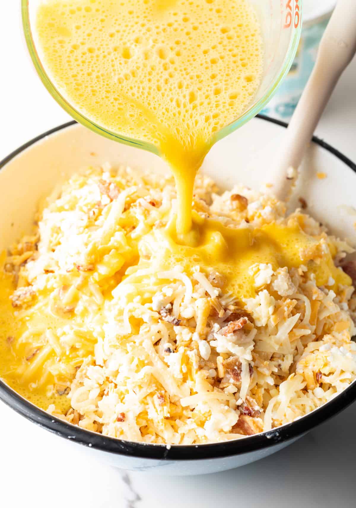 Pouring egg mixture from a glass measuring pitcher into a white bowl with thawed hash brown potatoes, cheese and bacon.