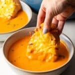 A hand holding cheesy bread that is being dipped into a bowl of roasted red pepper soup.