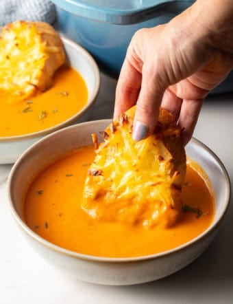 A hand holding cheesy bread that is being dipped into a bowl of roasted red pepper soup.