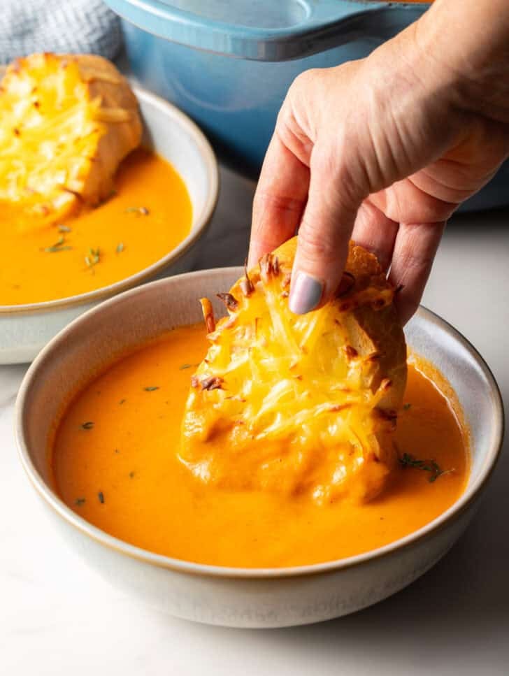 A hand holding cheesy bread that is being dipped into a bowl of roasted red pepper soup.
