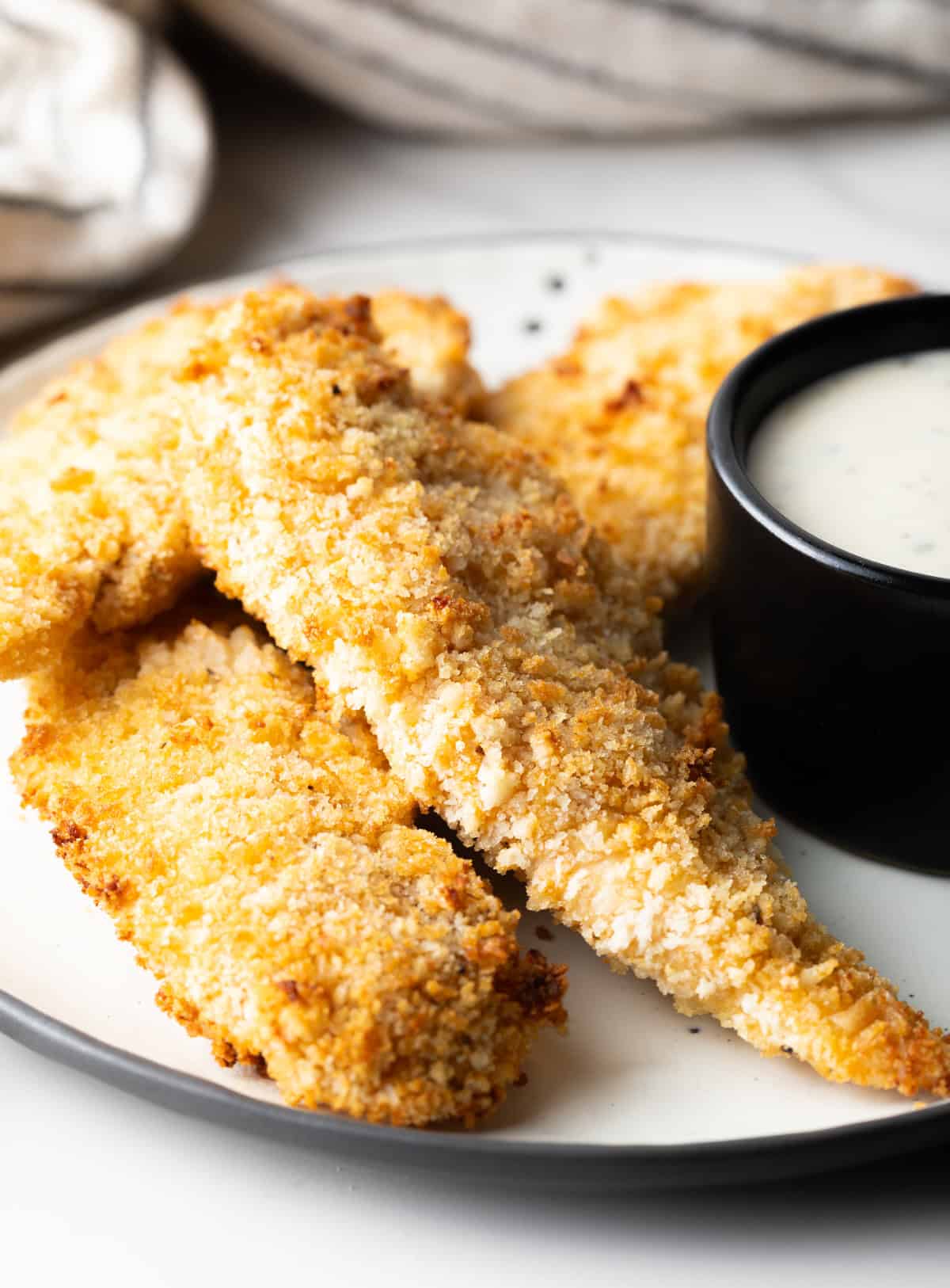 A side view of a white plate filled with crispy air fryer chicken tenders with a ramekin of dipping sauce on the side.