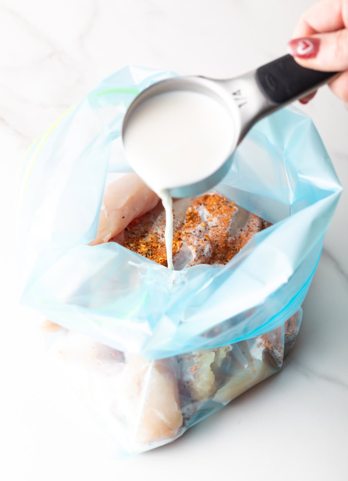 A ziplock bag filled with raw chicken tenders and seasonings, with buttermilk being poured into the bag with a measuring cup.
