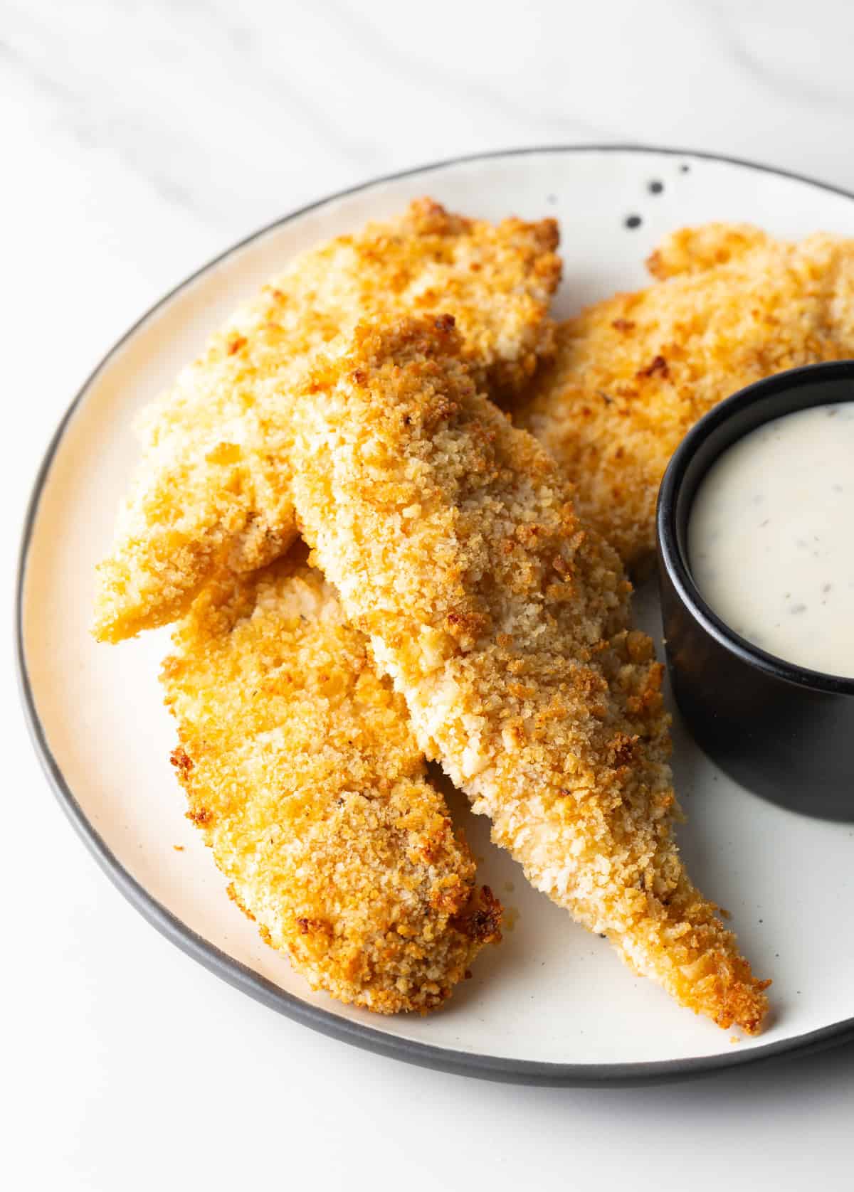 A plate of crispy air fryer chicken tenders next to a ramekin of white sauce.