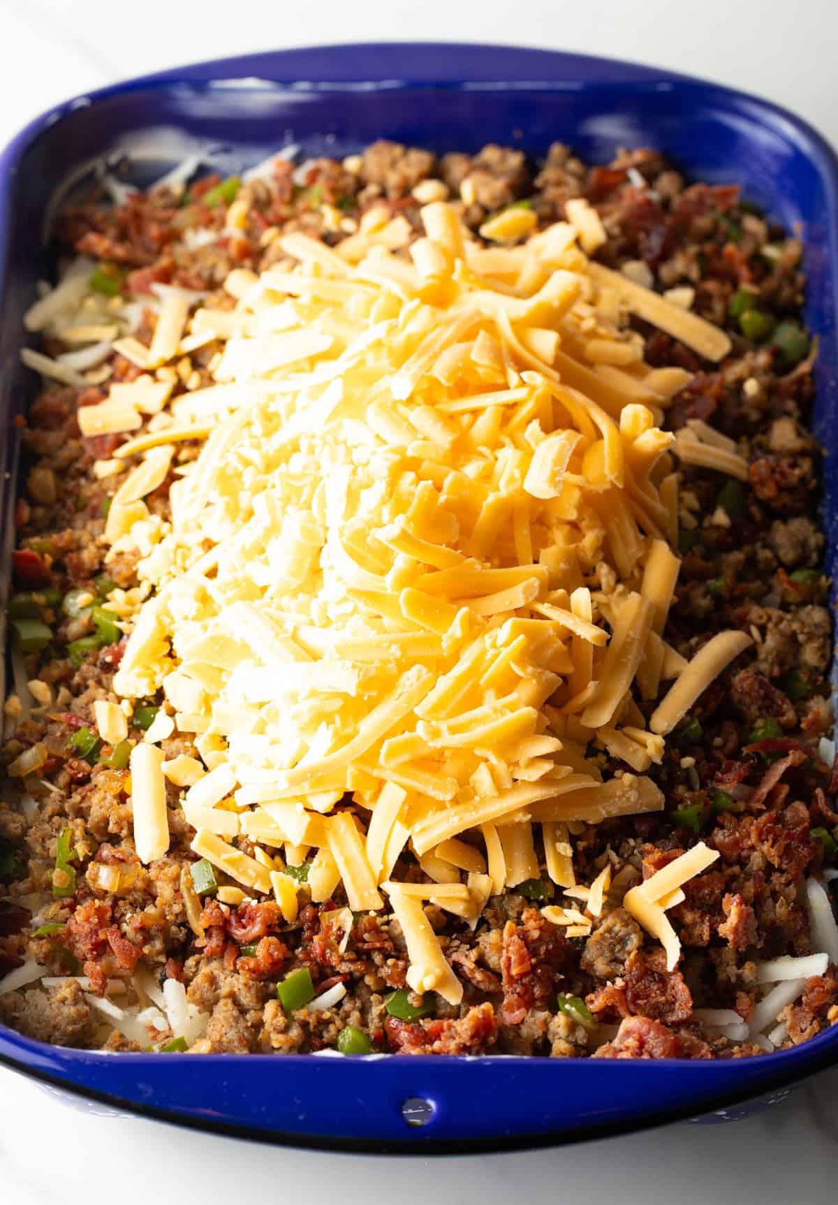 Shredded cheese is being added to a casserole dish filled with hash browns and sausage.