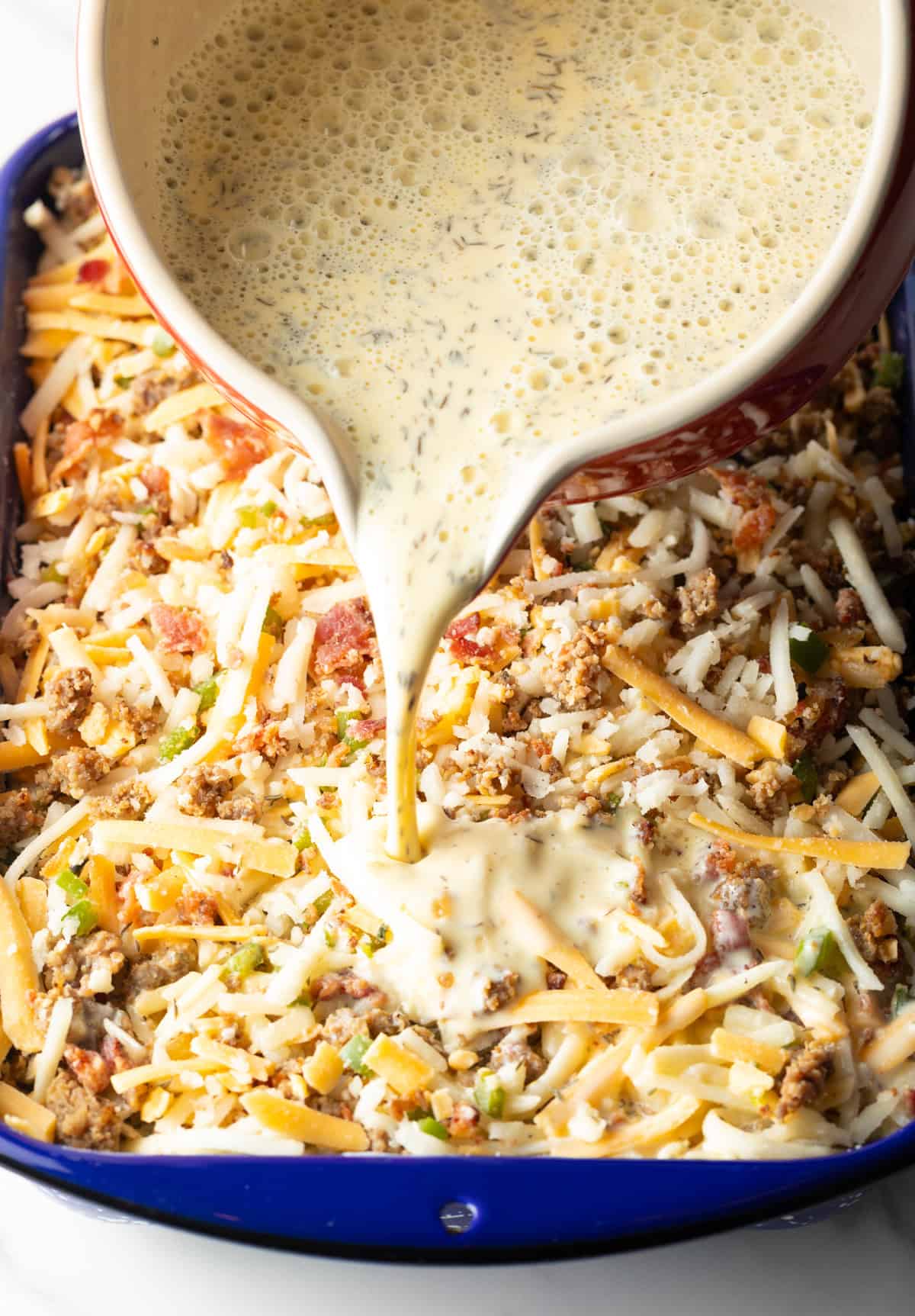An egg and cream mixture is being poured over ingredients for a hash brown breakfast casserole.